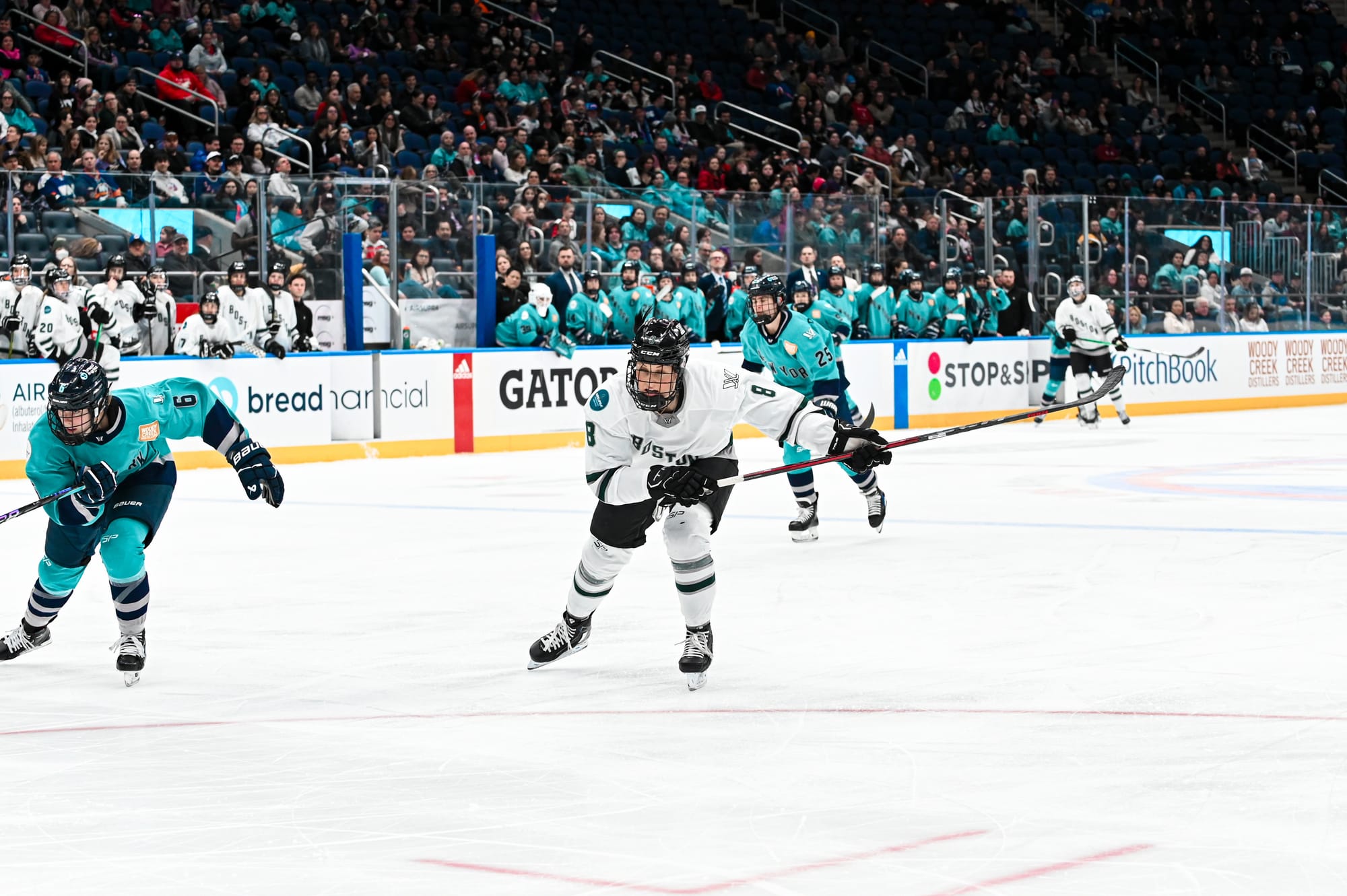Lexie Adzija hunts the puck. She is hunched over while skating hard. Her stick is out to her left. She is wearing a white away uniform.