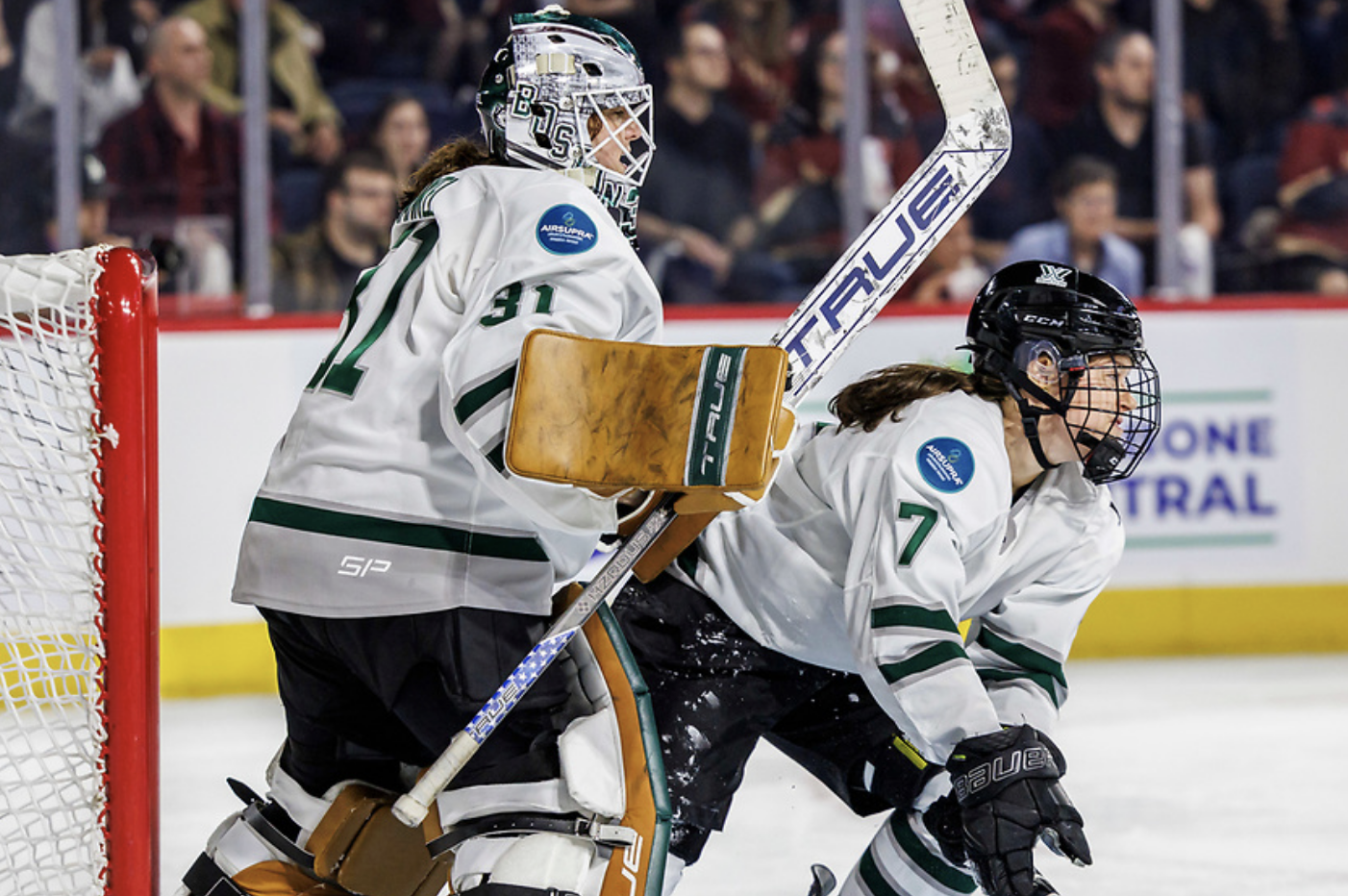 Frankel is standing, preparing to make a save, while Morin is right in front of her, hunched over trying to defend. They are both in white away uniforms.
