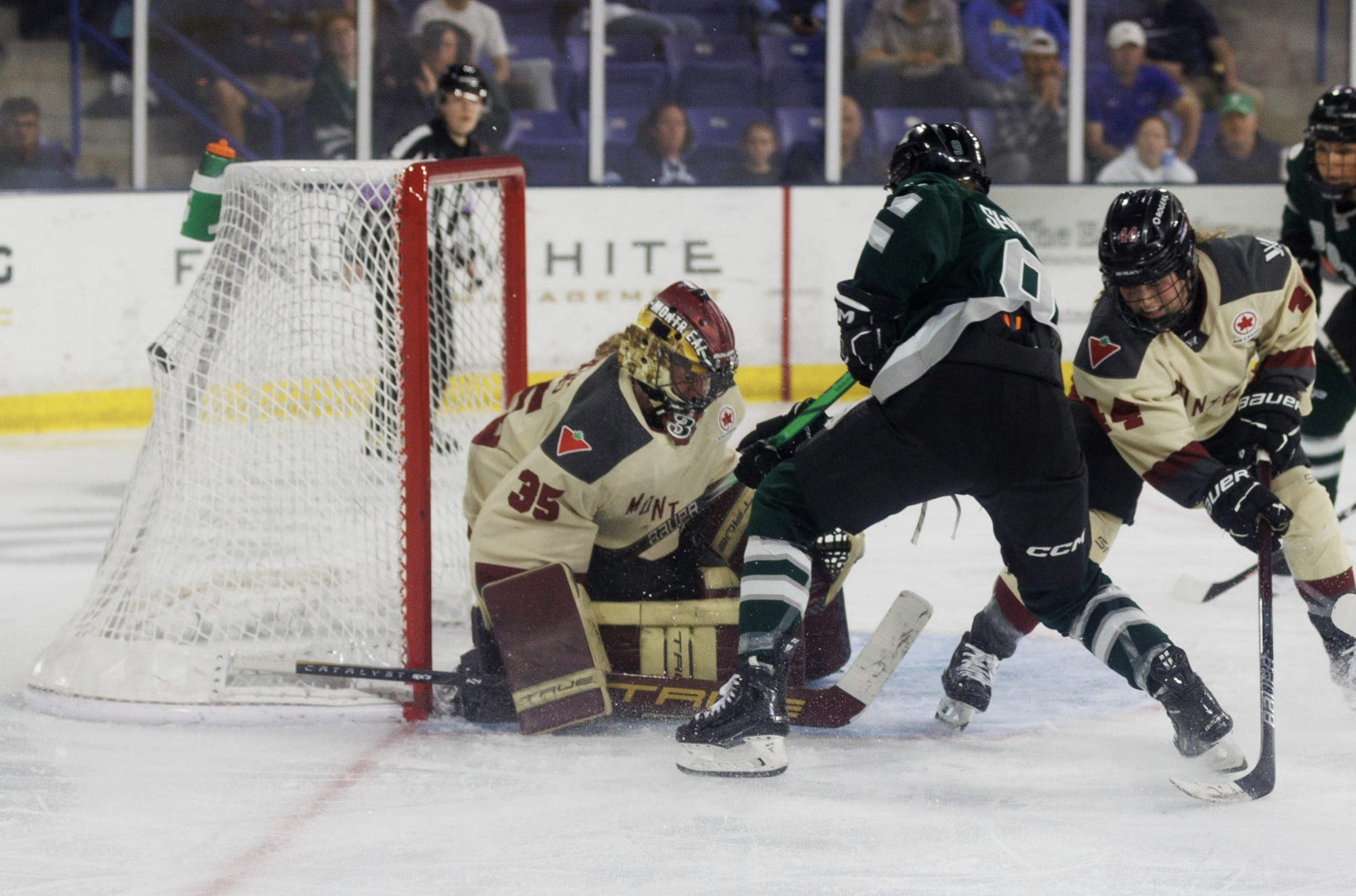 Desbiens is on her knees while Shirley is upright right in front of her crease. The puck has just hit the back of the net. Shirley is in a green home uniform, while Desbiens is in a cream away uniform.