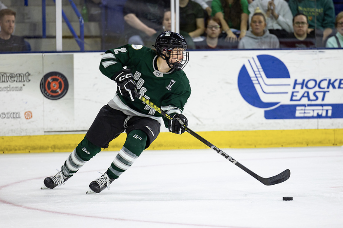 Brown turns to skate forward with the puck. She is looking up ice while wearing a green home uniform.
