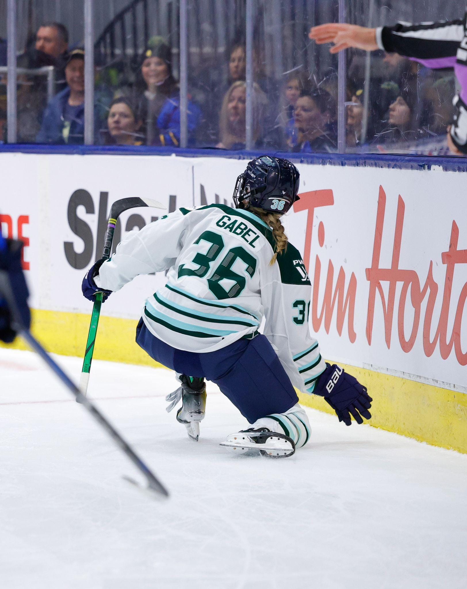 A from-behind view as Gabel drops down to one knee and reaches her right hand down towards the ice in celebration. She is wearing a white away uniform.