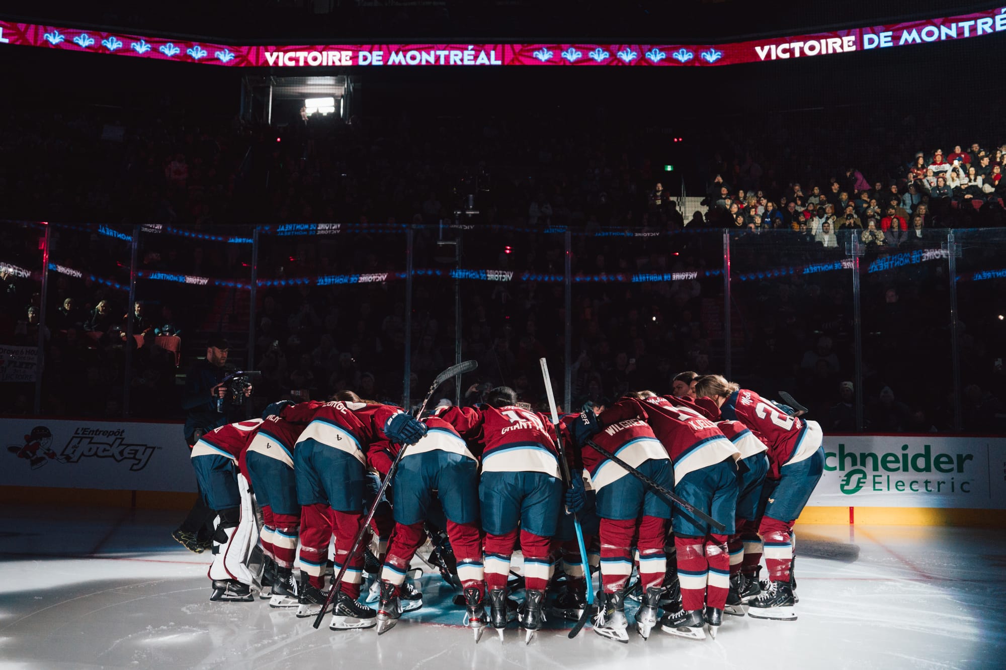 Victoire players stand close together. They're all hunched over, leaning into the huddle in front of one of the goals. They are wearing maroon home uniforms.