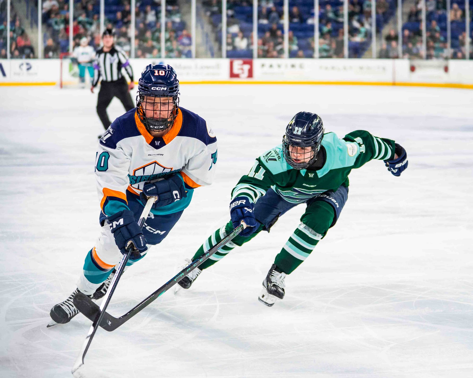 Fillier (left in white) looks down at the puck as she skates with it in front of her. Müller (right in green) is leaning forward as she goes for the poke check. The puck is out of the frame.