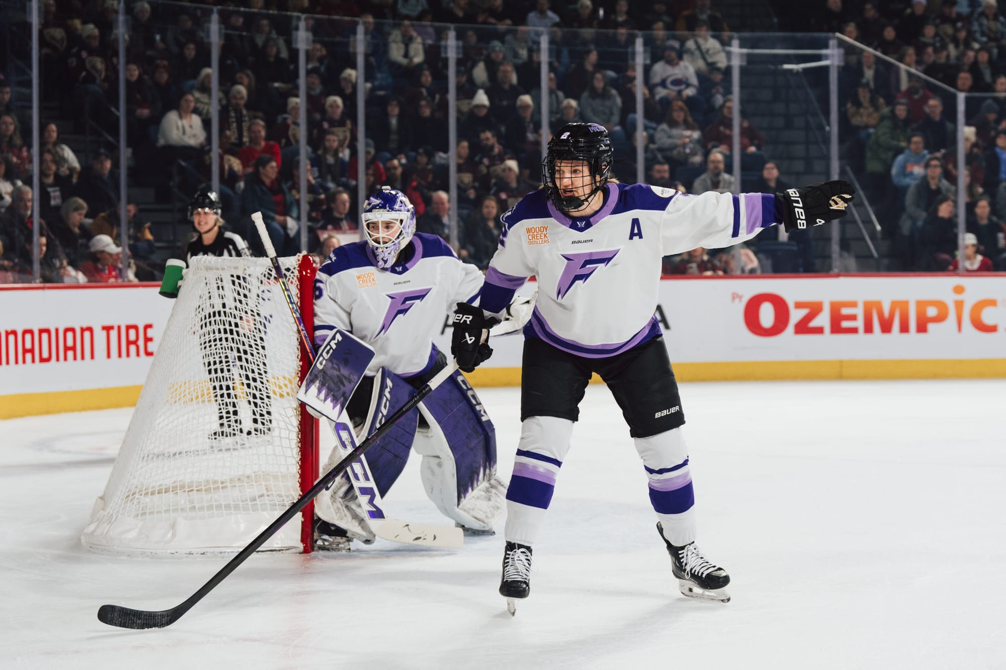Stecklein stands by the goal crease and points to her left, with her goaltender behind her. She is wearing a white away uniform.