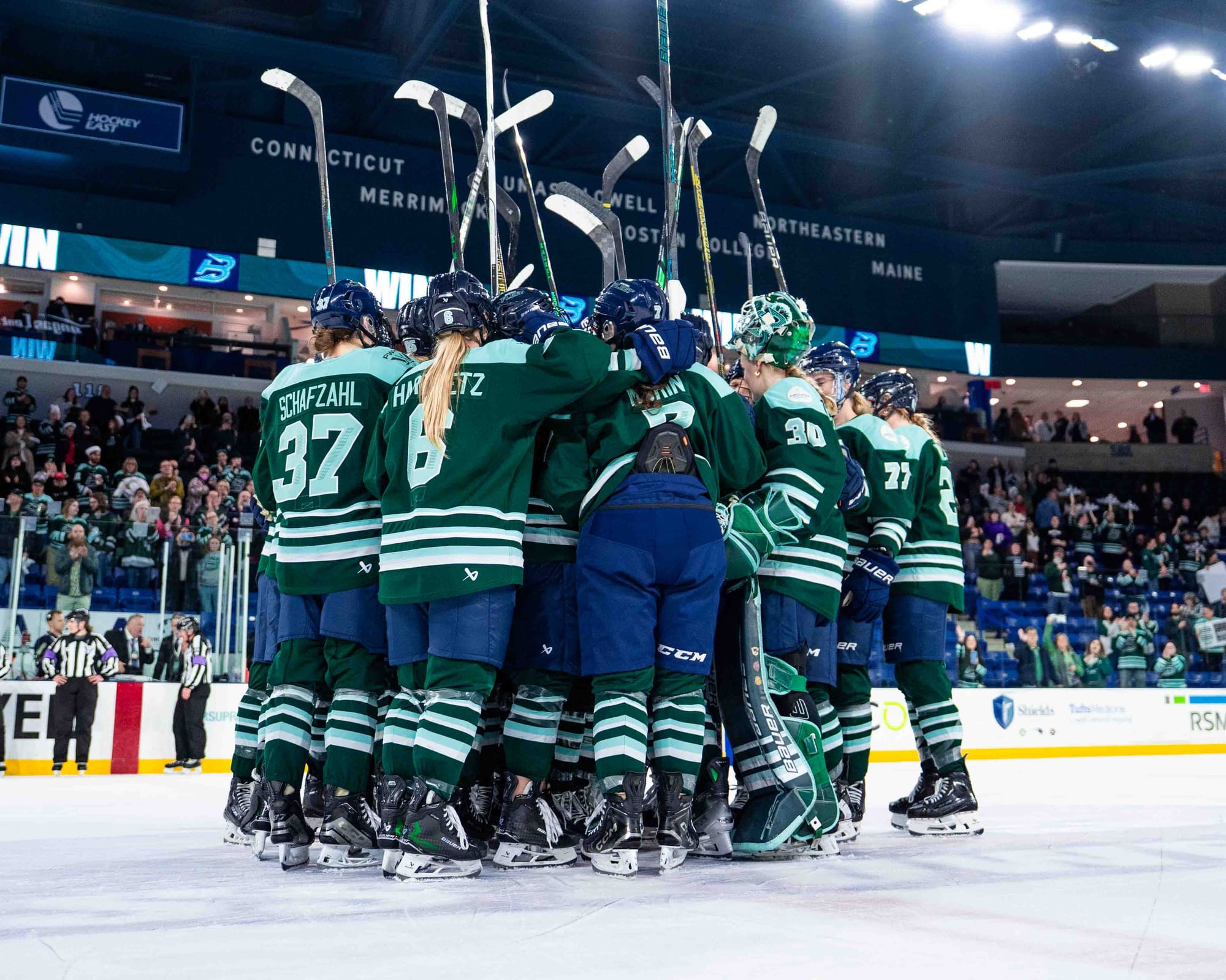 Fleet players raise their sticks and celebrate with a tight group hug at center ice. They are wearing green home uniforms.