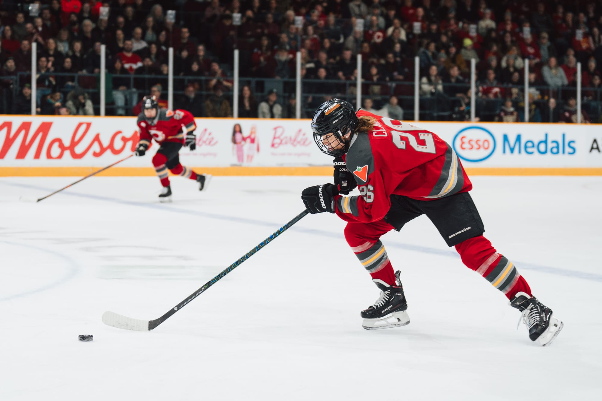 Clark pushes the puck in front of her as she skates hard toward the center ice line. One of her teammates is out of focus in the background, providing a passing option. They are wearing red home uniforms.