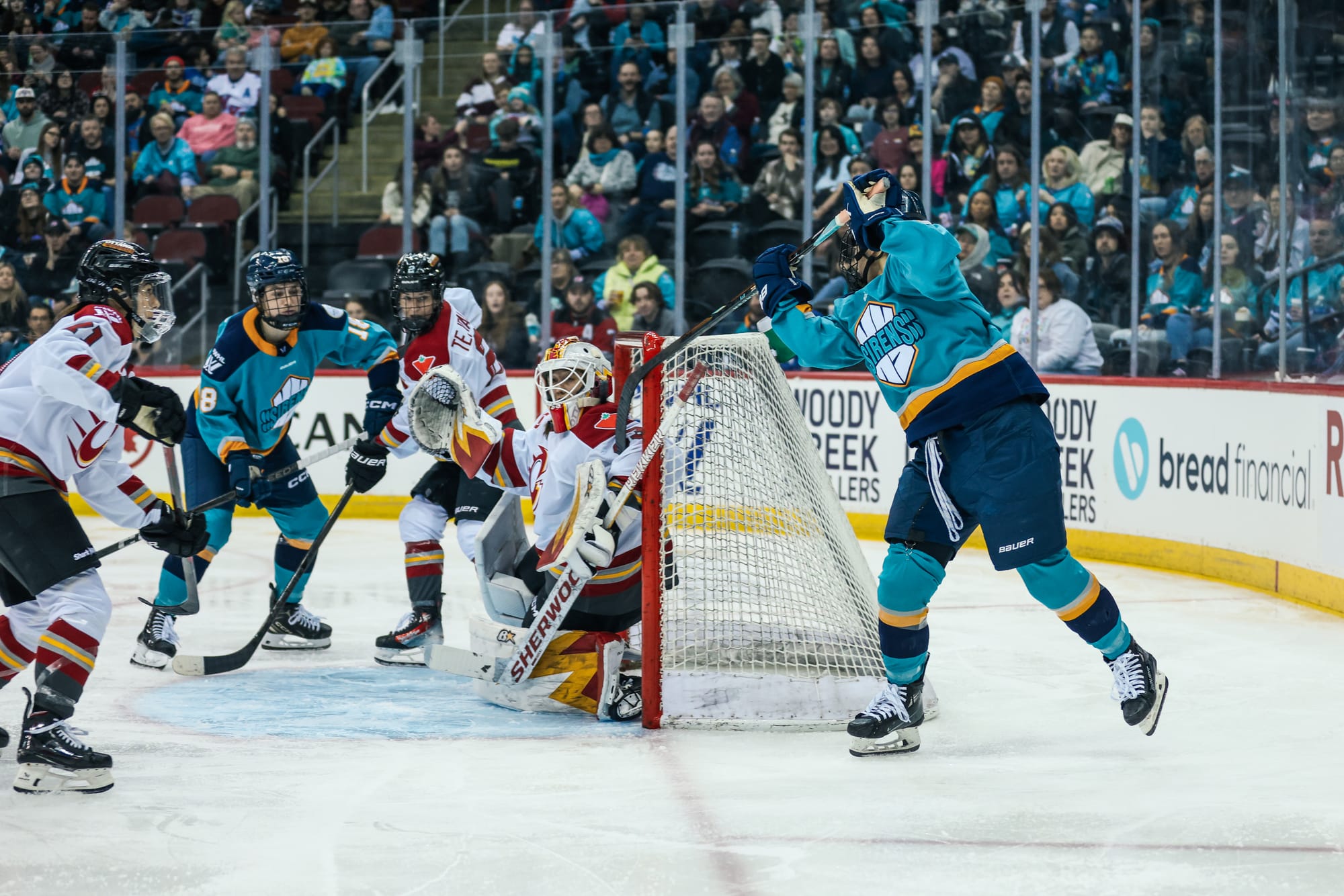 Roque stands next to the net, behind the goal line as she swings the blade of her stick, which has the puck on it, around the front from about shoulder height and stuffs the puck in. She is wearing a teal home uniform. There are other Sirens and Ottawa players out in front of the crease.