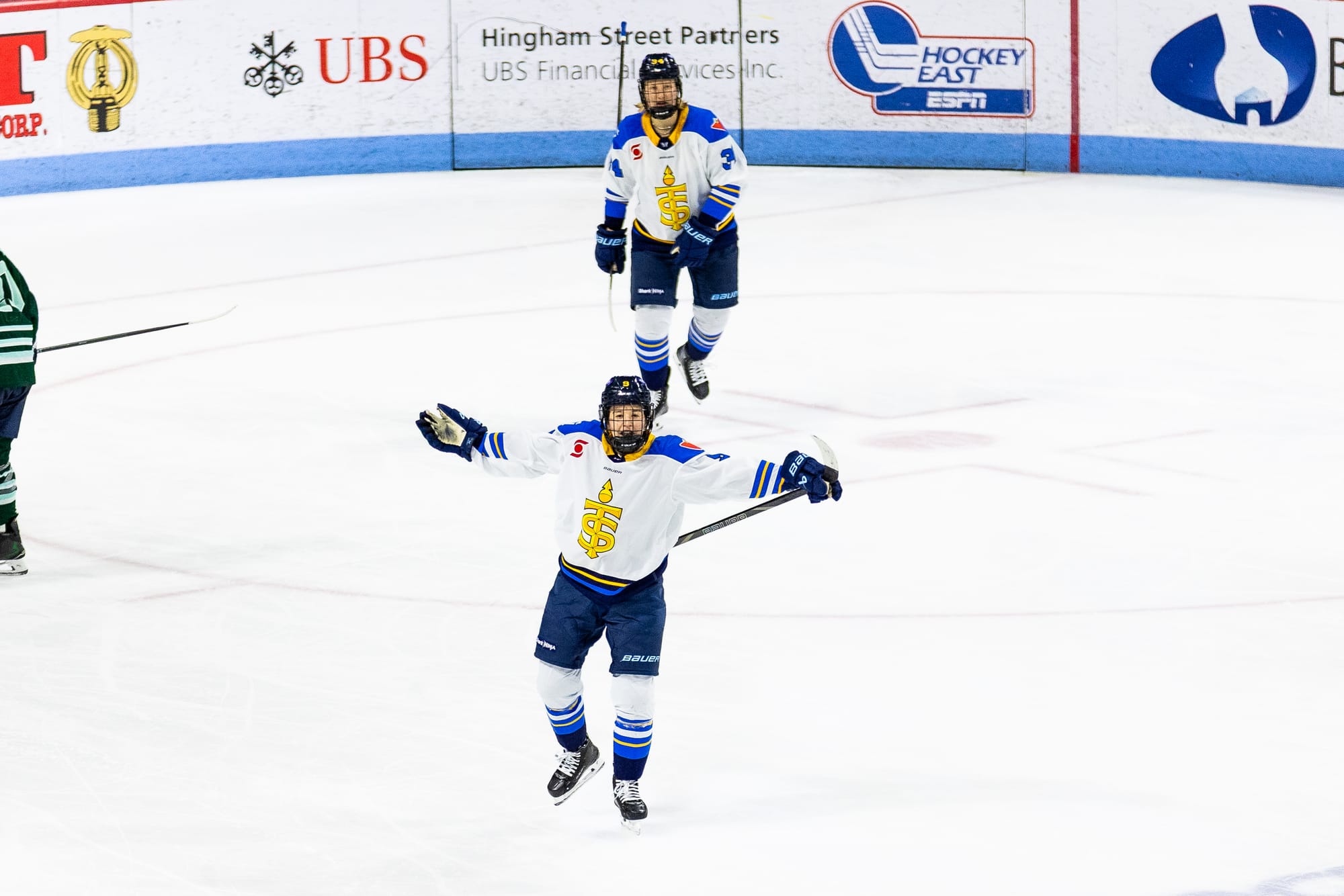 Watts raises her arms out to her sides, smiles, and yells in celebration. Hannah Miller is skating behind her. They are wearing white away jerseys.