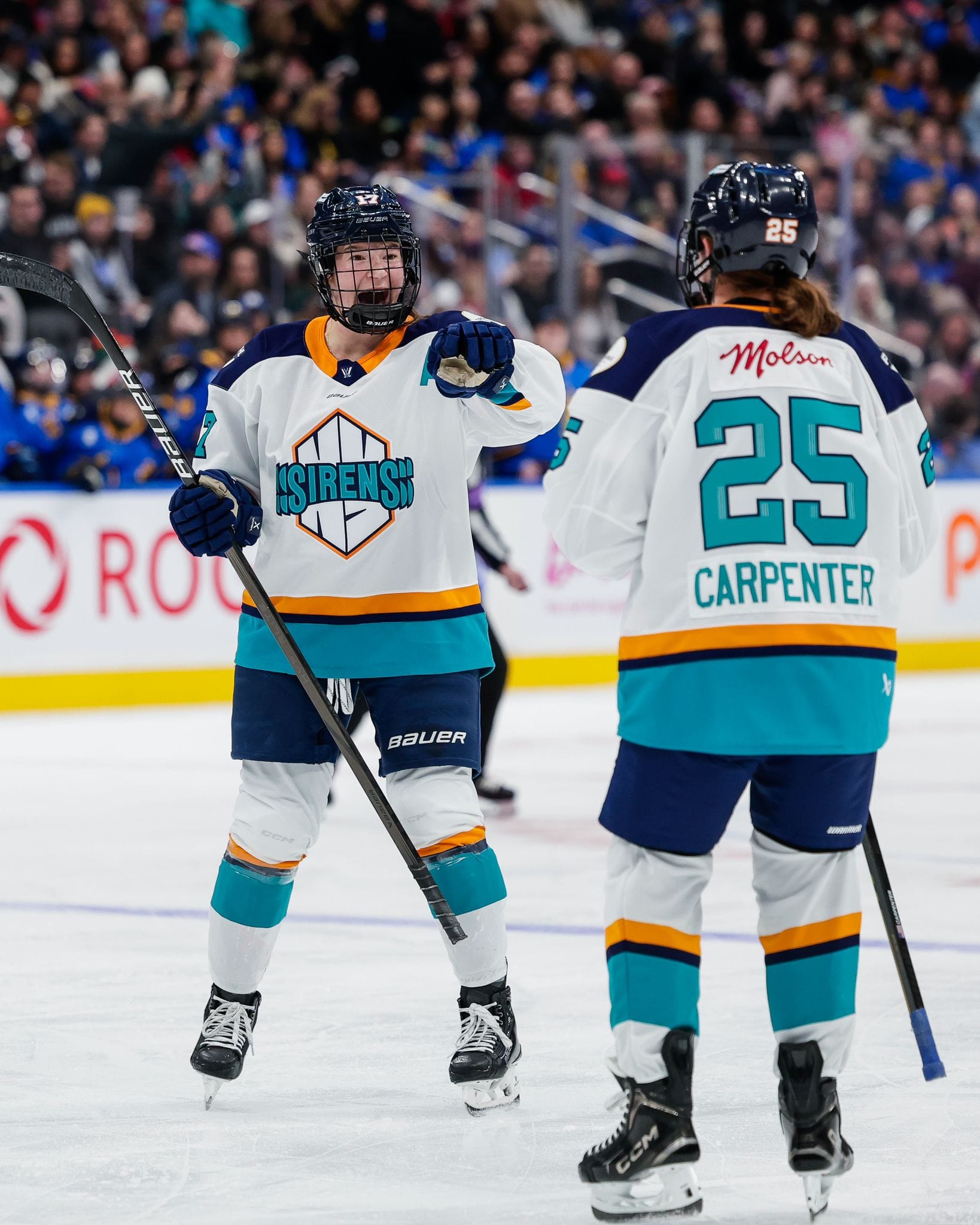 Shelton smiles and points at Carpenter, who is skating toward her. They are near the blueline and wearing white away uniforms.