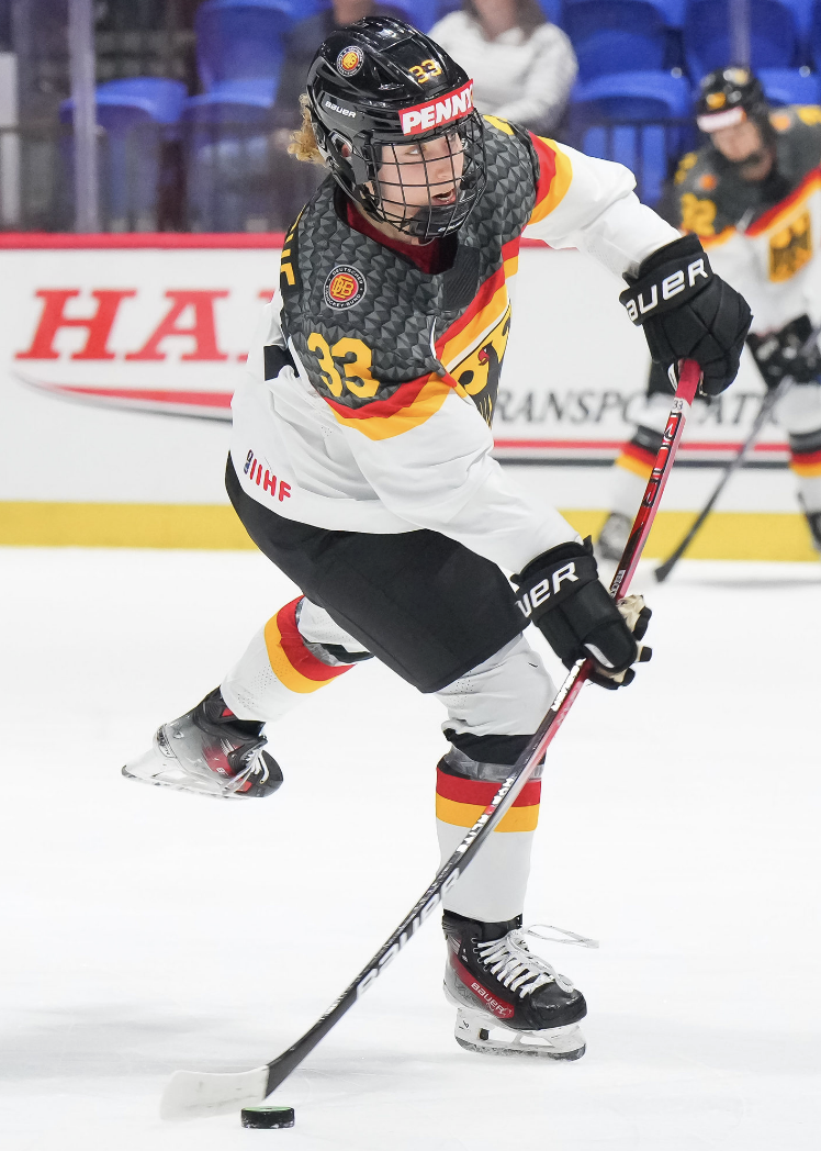 Welcke flexs her stick as she prepares to take a shot. She is looking ahead at the goal and wearing a white Germany uniform.