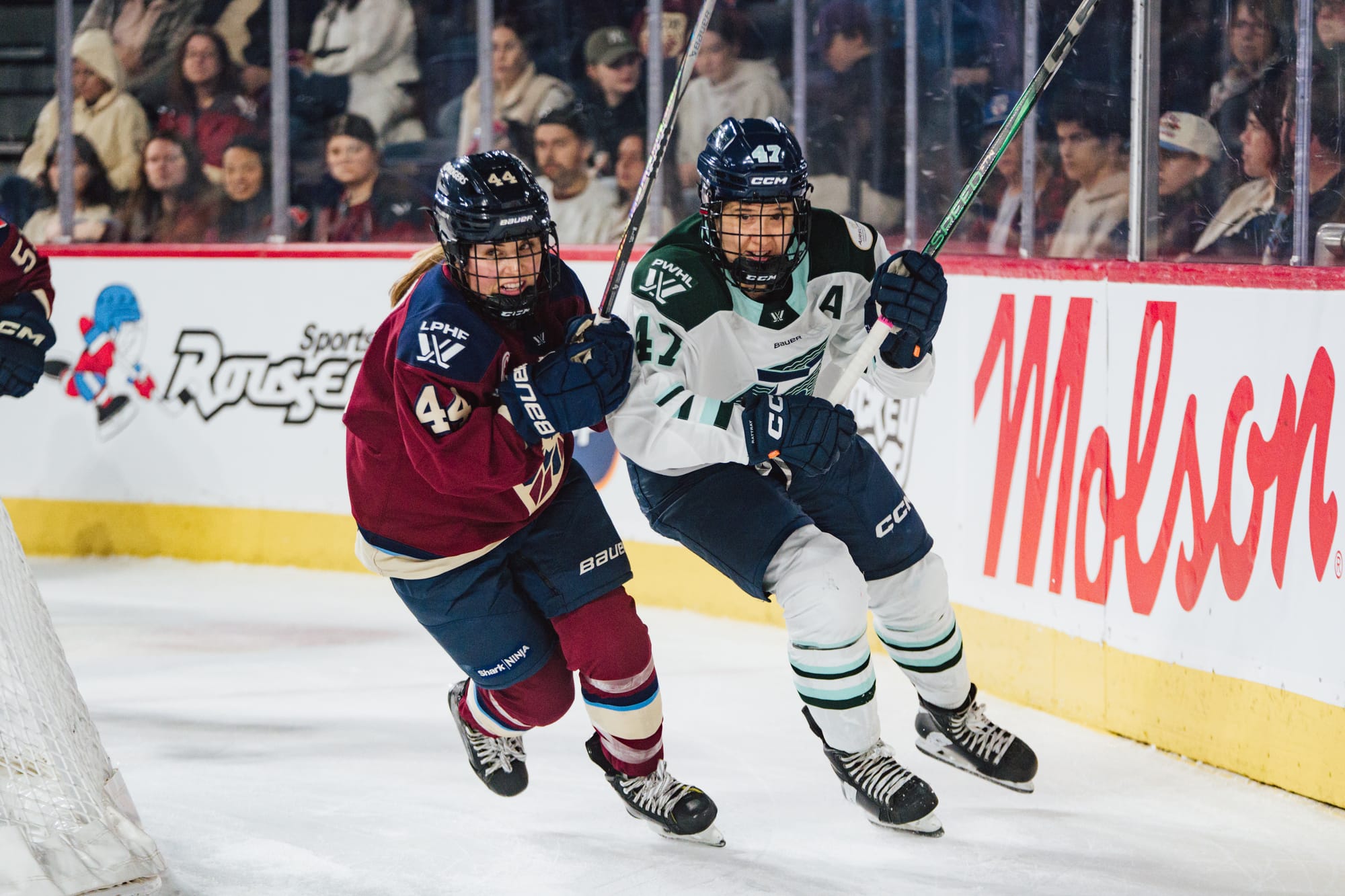 Boulier (left in maroon) and Rattray (right in white) skate behind the net in pursuit of the puck. Both have their sticks in the air as they skate around the corner.