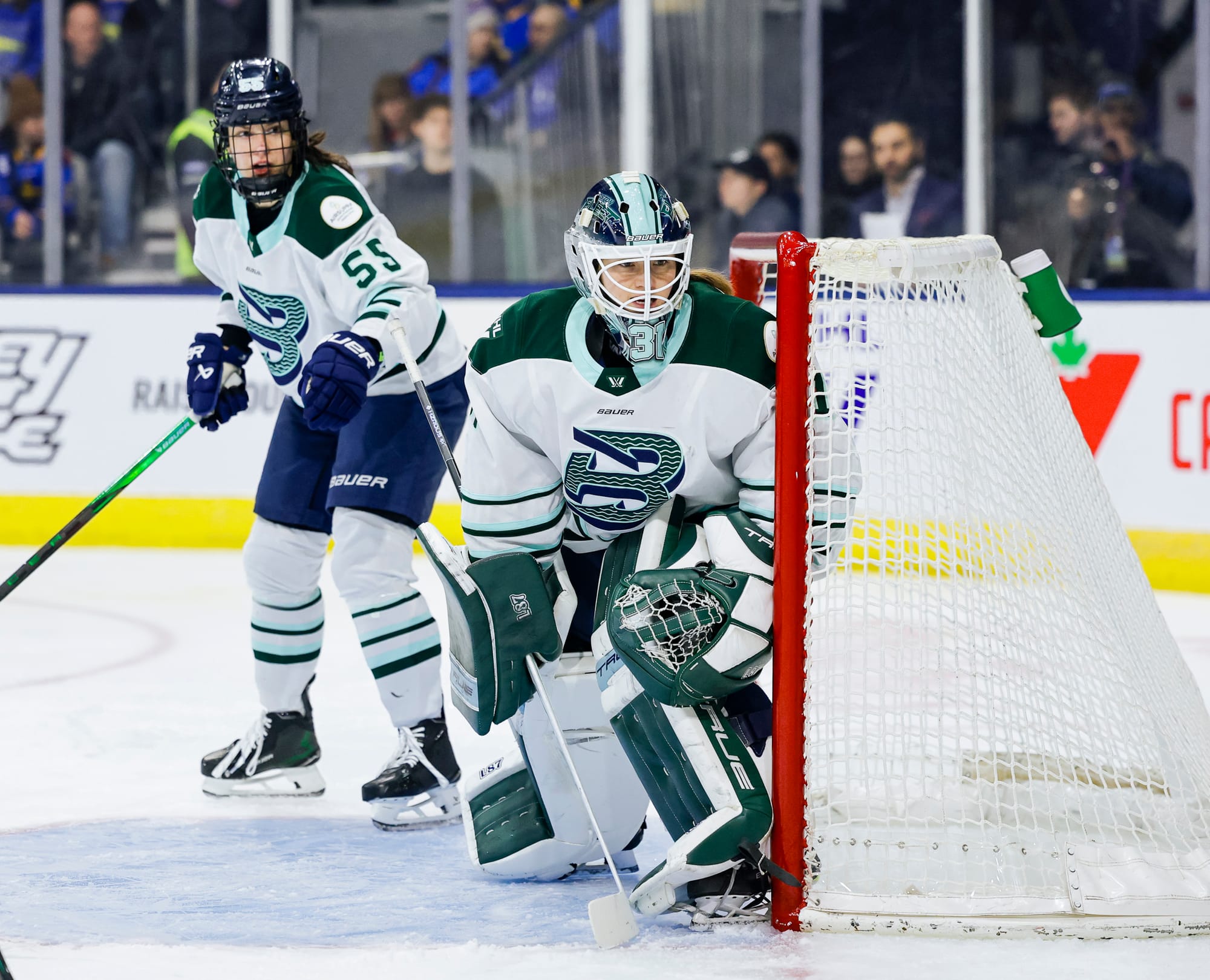 Pejšová stands just outside the crease to Frankel's right. Frankel is on the left side of the net, watching a play off to her left behind the goal line. They are both wearing white away uniforms.