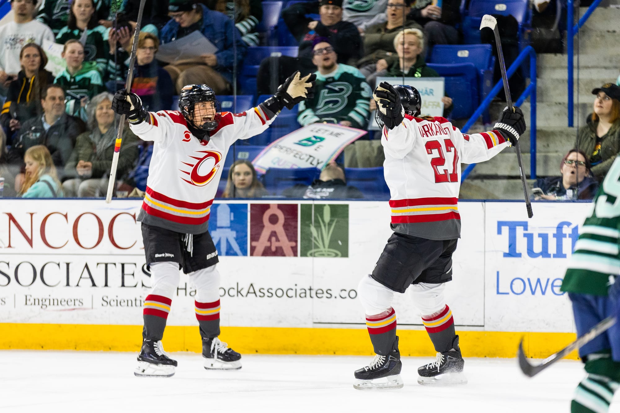 Darkangelo and her teammate skate toward each other with wide open arms. Darkangelo's back is toward the camera, but her teammate is smiling. They are both wearing white away uniforms.
