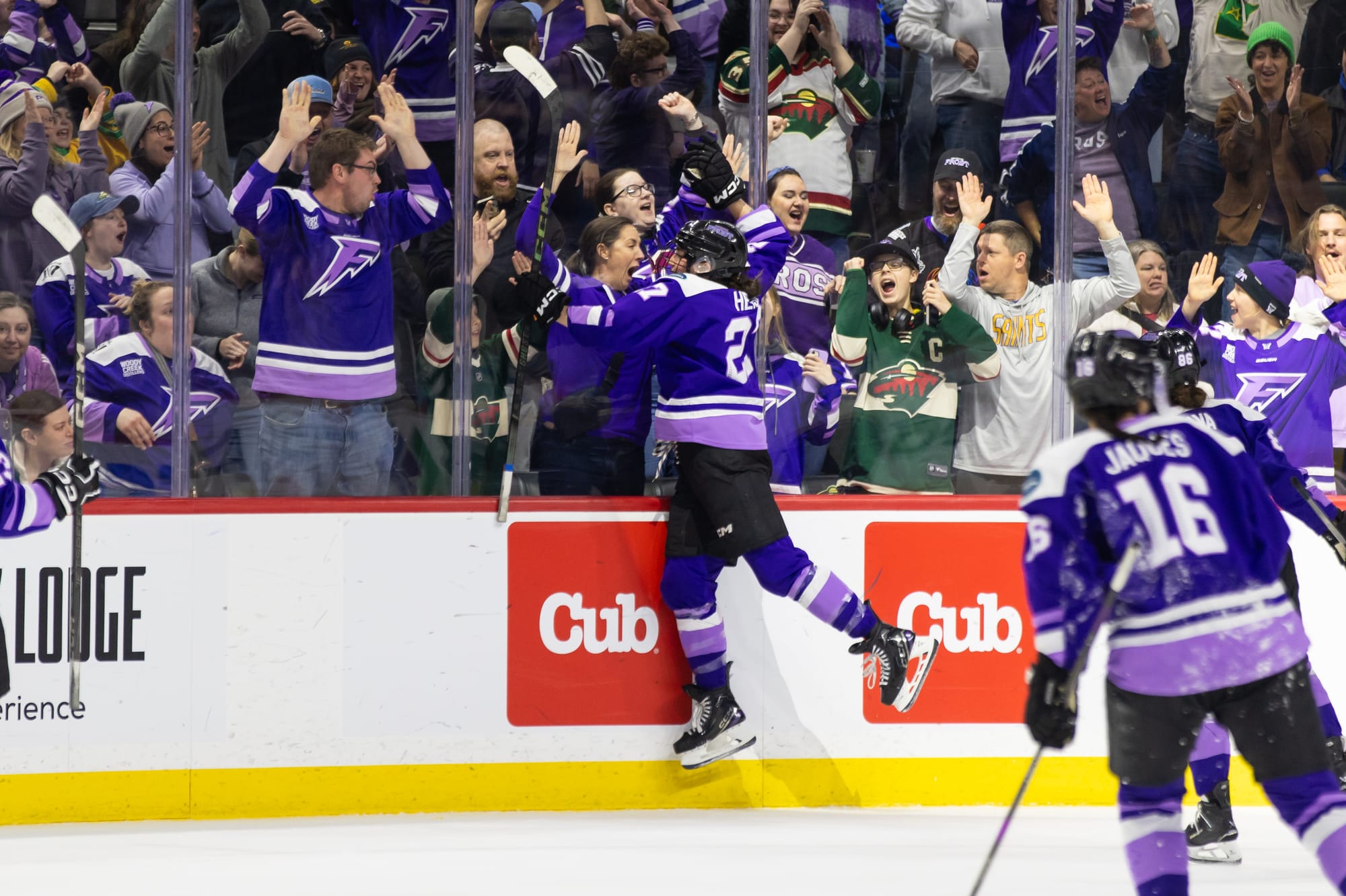 Heise jumps into the boards to celebrate with fans, while her teammates skate toward her for a hug. The players are all wearing purple home uniforms.