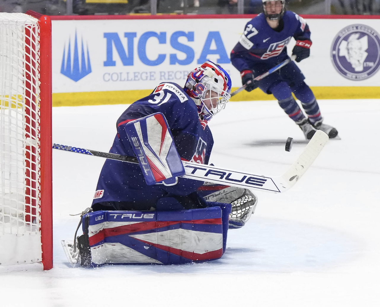 Frankel looks down as she prepares to make a save from her knees.She is wearing a blue uniform and her red/white/blue USA pads.