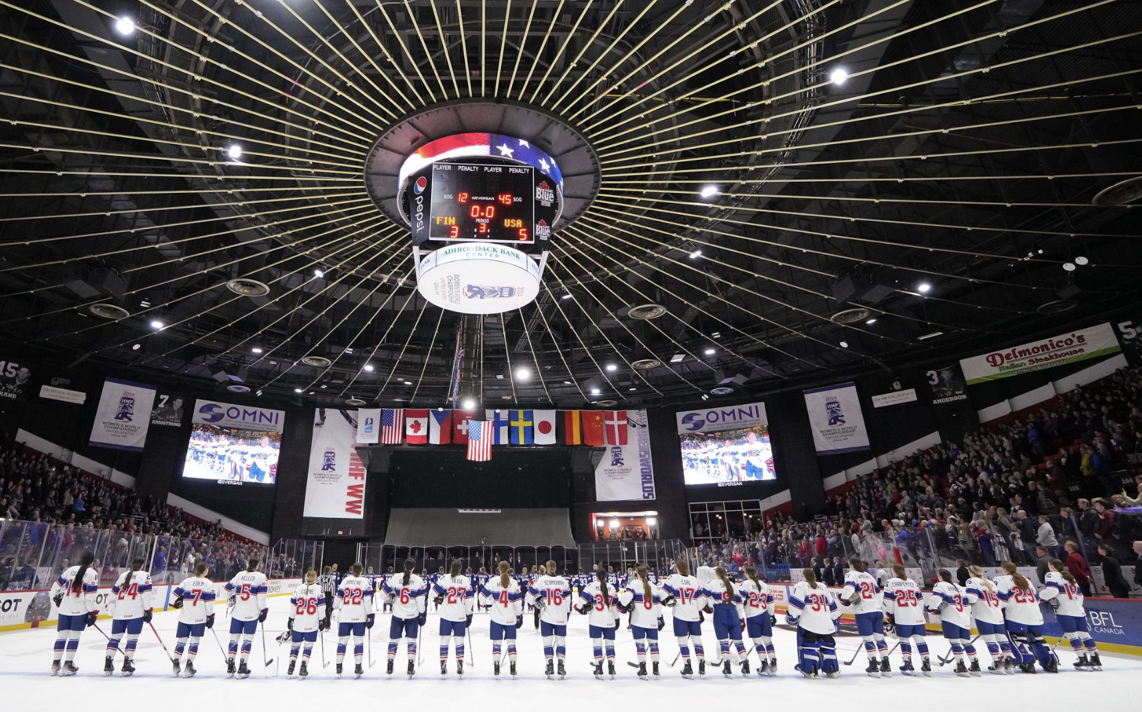 Team USA stands shoulder-to-shoulder along the blue line and looks at the flag as the anthem plays. They are wearing white uniforms with a blue stripe in the middle.
