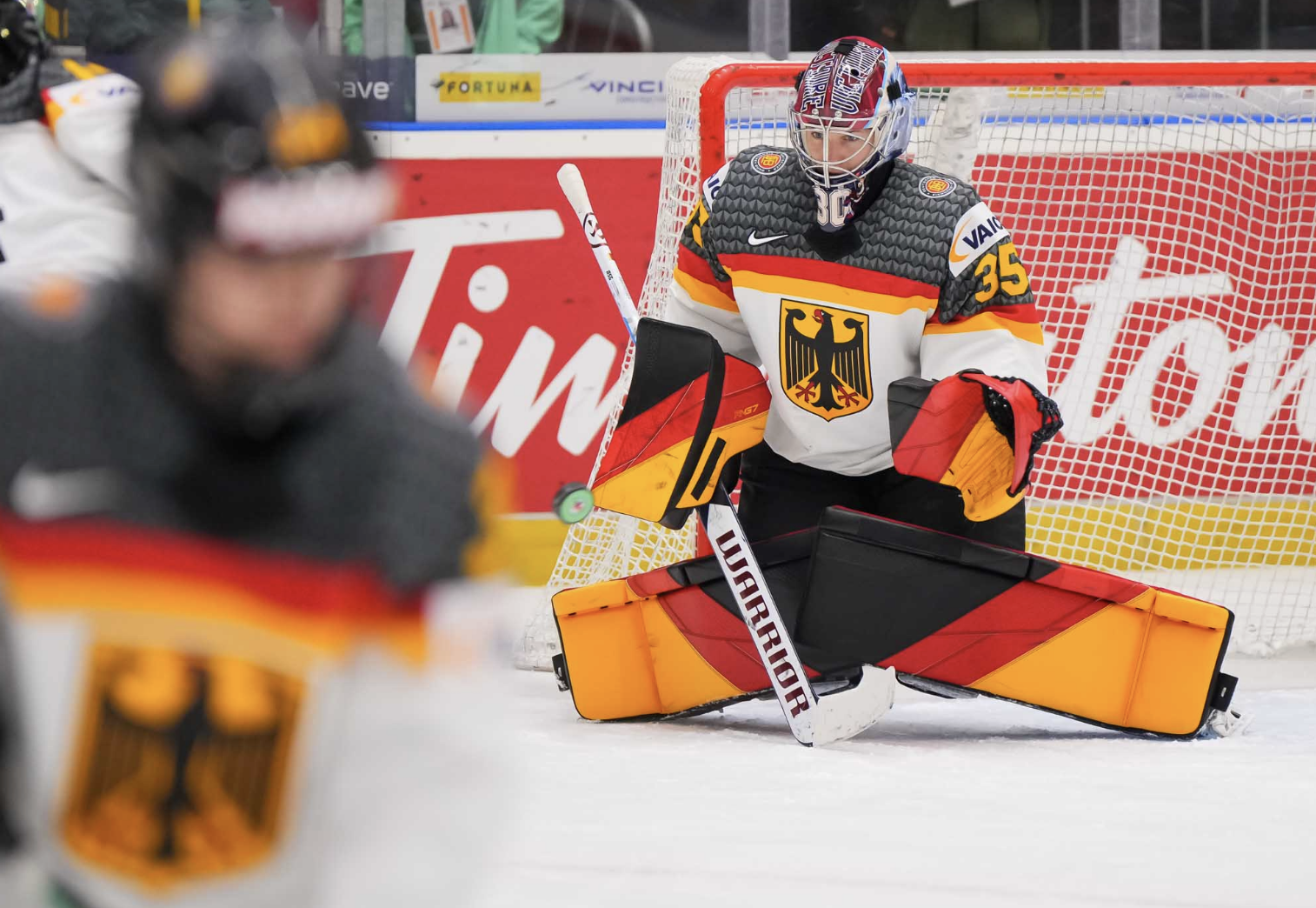 Abstreiter drops to the butterfly position to make a save during warmups. She is wearing a white uniforms and black/red/orange gear that is striped to look like the German flag.