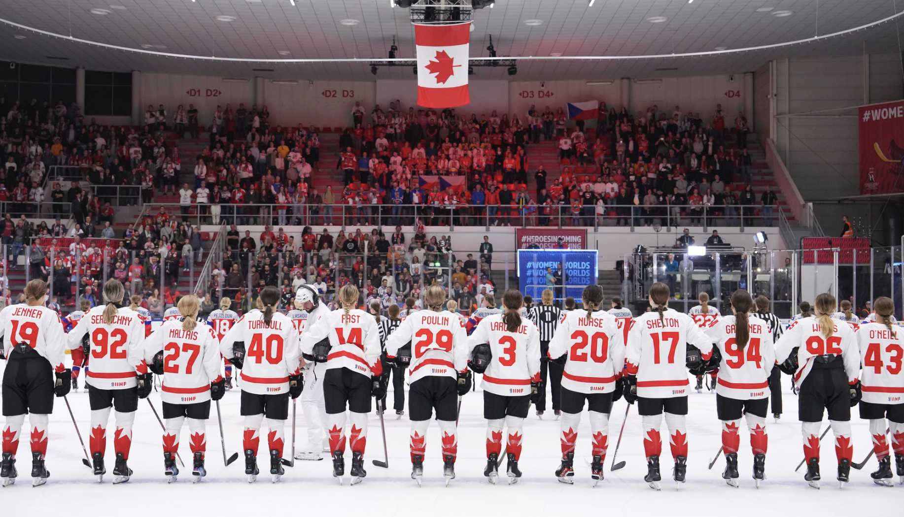 Canadian players stand shoulder to should along the blue line while the national anthem plays. They are wearing white uniforms.