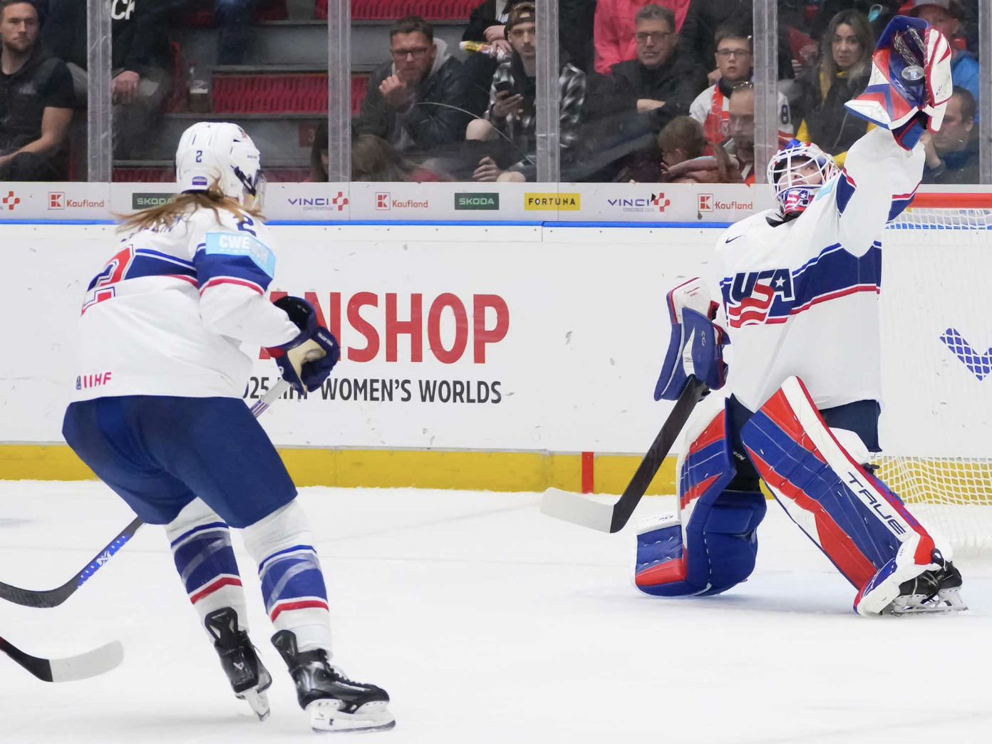 Frankel stands in her crease, with her left arm raised high above her head and the puck in her glove. Stecklein is skating in from the left, prepared to defend if necessary. They are wearing white uniforms.