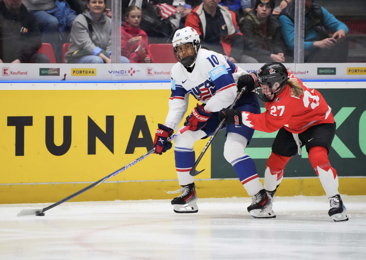 Edwards (left in white) looks to her left as she skates with the puck, preparing to pass it. Maltais (right in red) is pushing her, trying to knock her off the puck.
