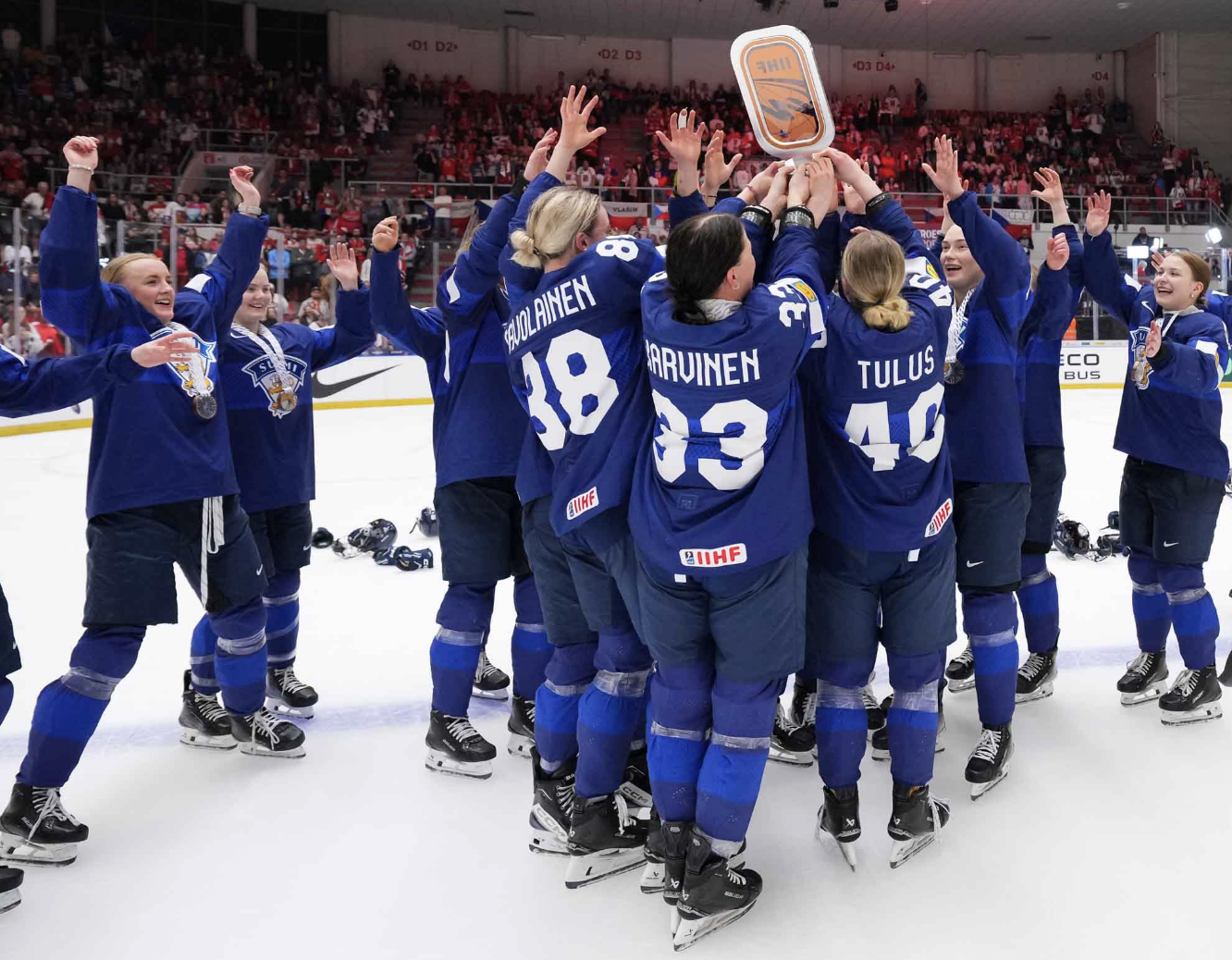 Team Finland fully surrounds their captain and tries to touch the bronze medal trophy she's hositing. They are all wearing blue uniforms. 