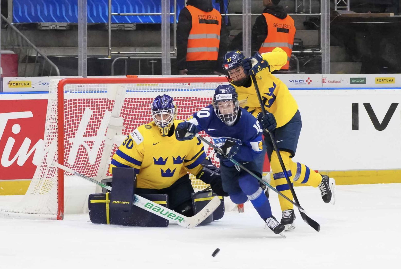 Söderberg is on her knees, preparing to make a save as the puck comes toward her. Kjellbin is off to her left, battling for positioning with a Finland player.
