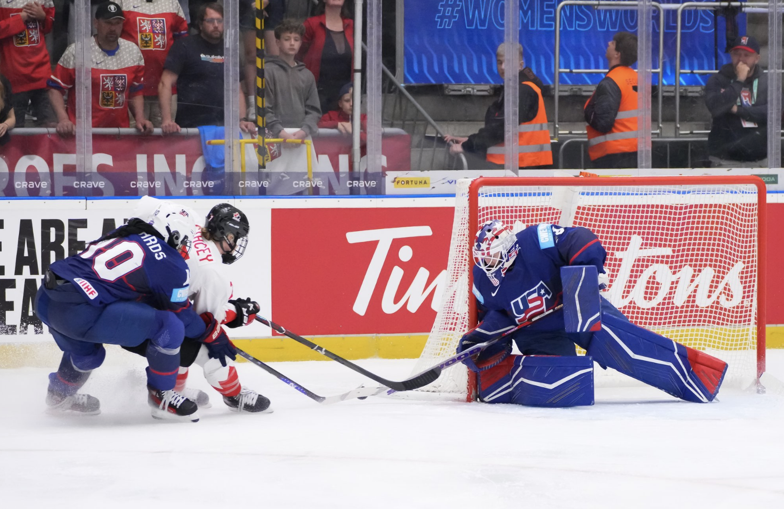 Phillips is on her knees, leaning on her right post as she pokes the puck away from Canada's Laura Stacey, who is being defended by Laila Edwards. The Americans are in blue uniforms, while Stacey is in white.