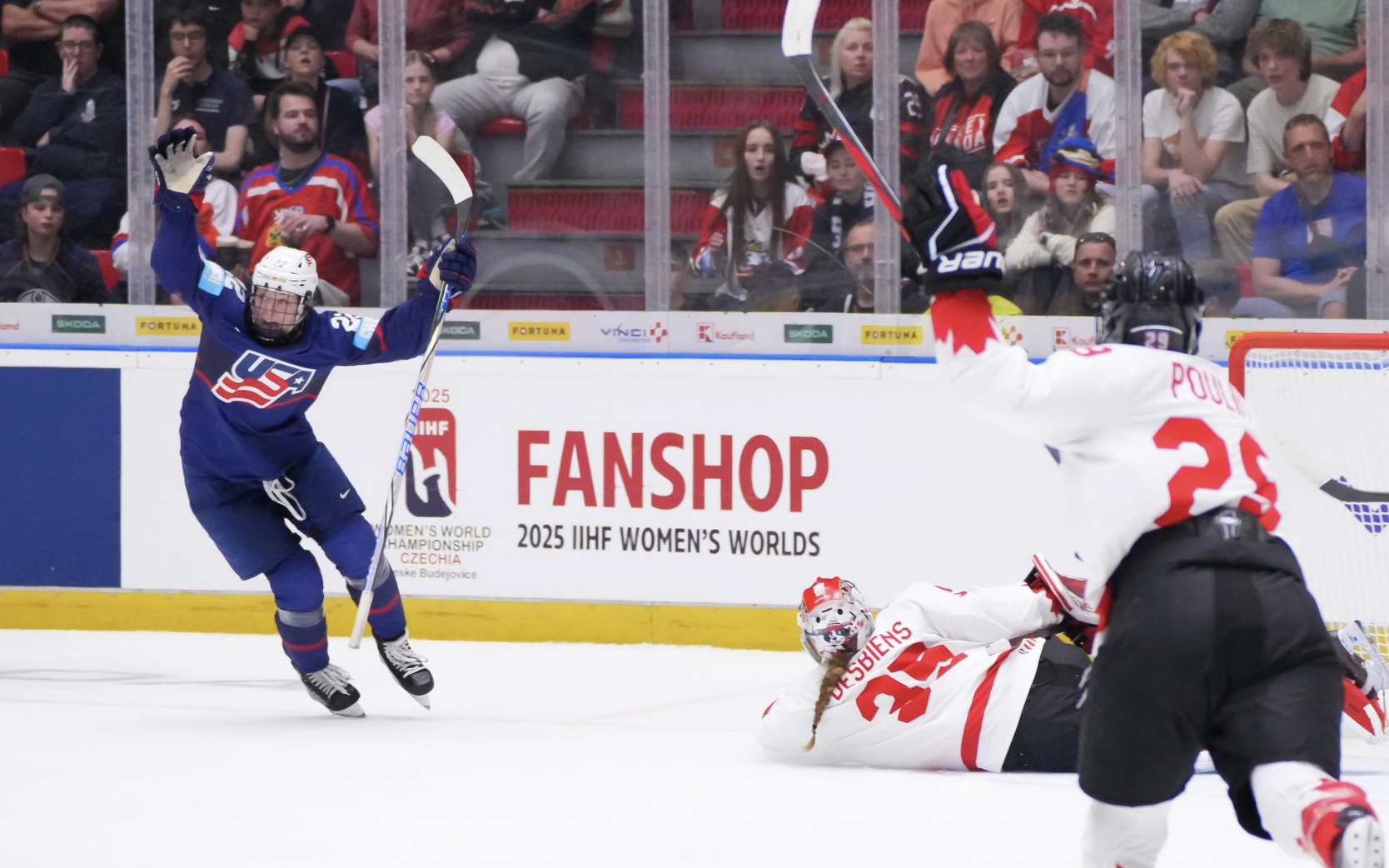 Janecke raises her arms as she starts to turn. She is wearing a blue uniform. Canada's Desbiens is laying on the ice, while Poulin is also skating into the frame (both are wearing white).