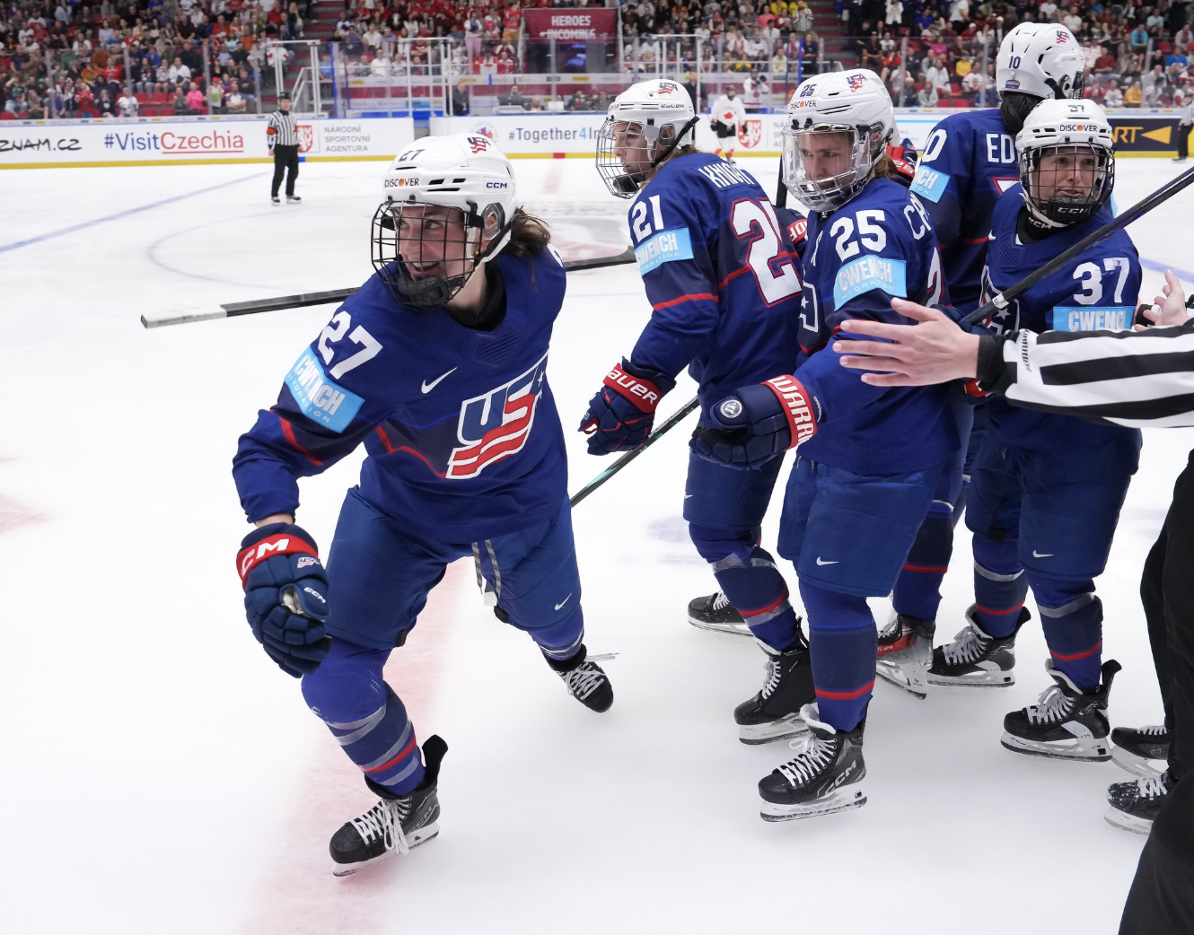 Heise smiles as she turns away from the group hug to skate toward the bench and down the handshake line. They are all wearing blue uniforms.