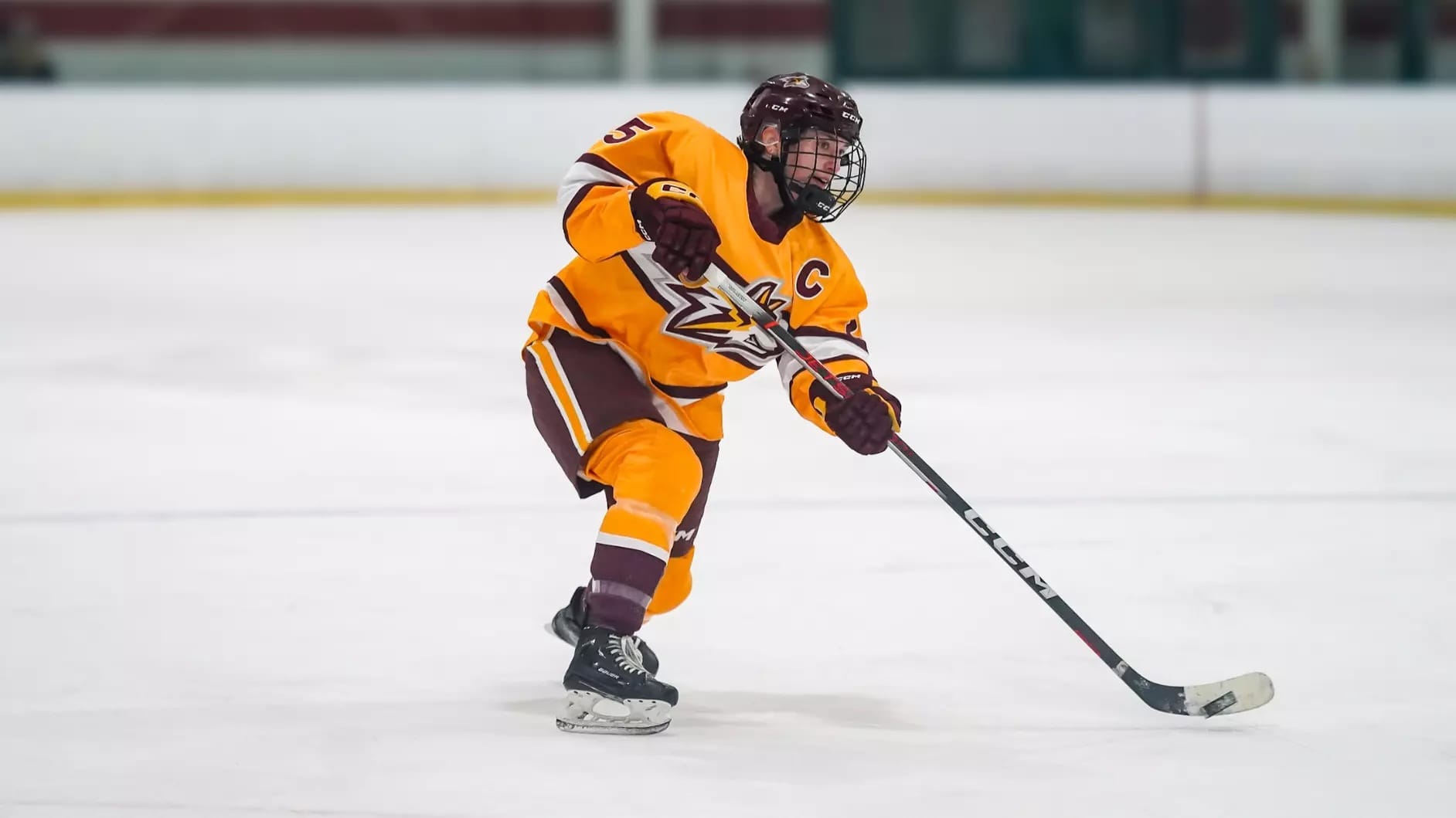 A left-handed hockey player in a gold uniform with maroon trim has the puck on her stick.