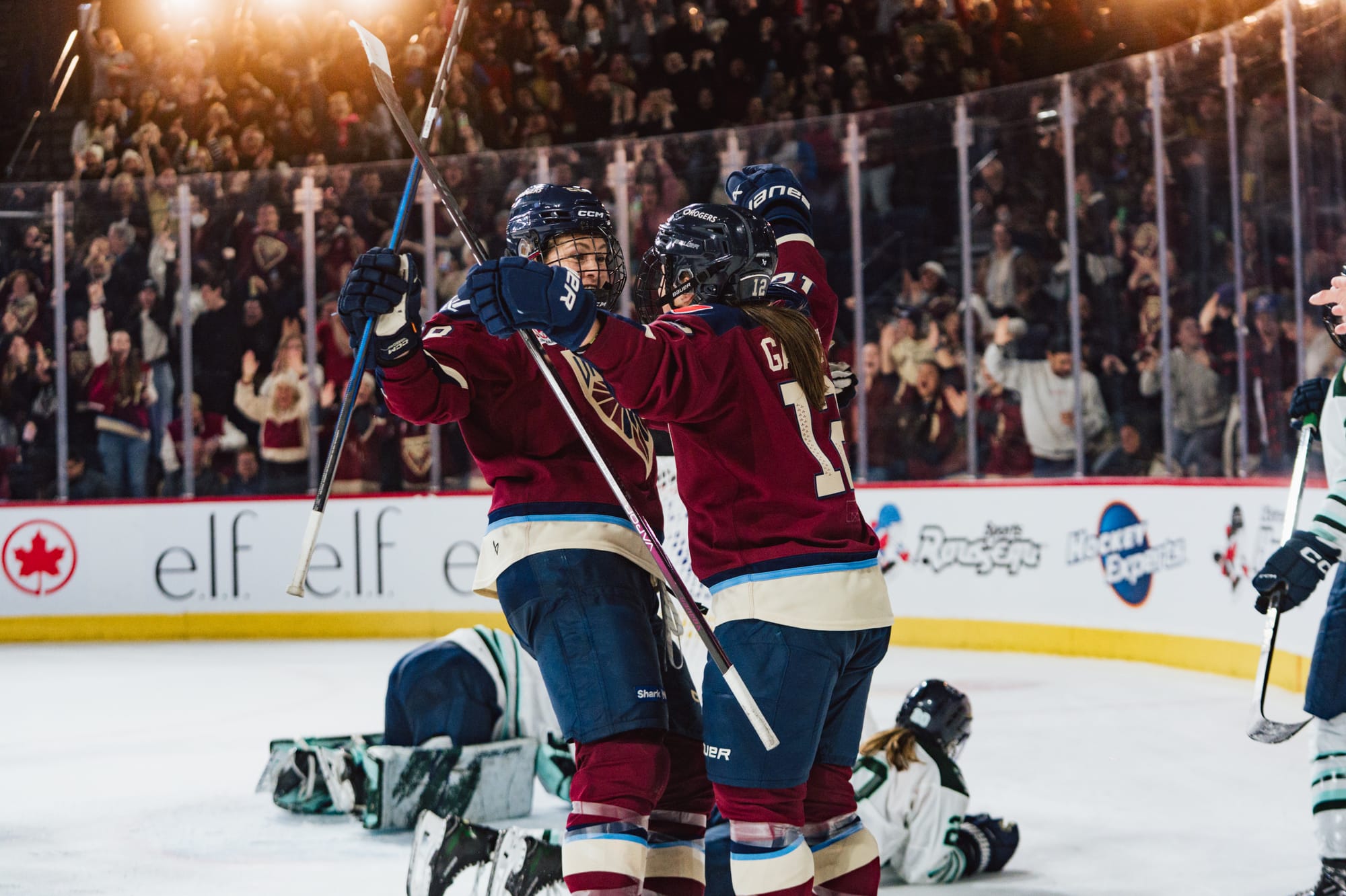 Gardiner and a teammate embrace to celebrate a goal, while the crowd cheers in the background. They are wearing maroon home uniforms.