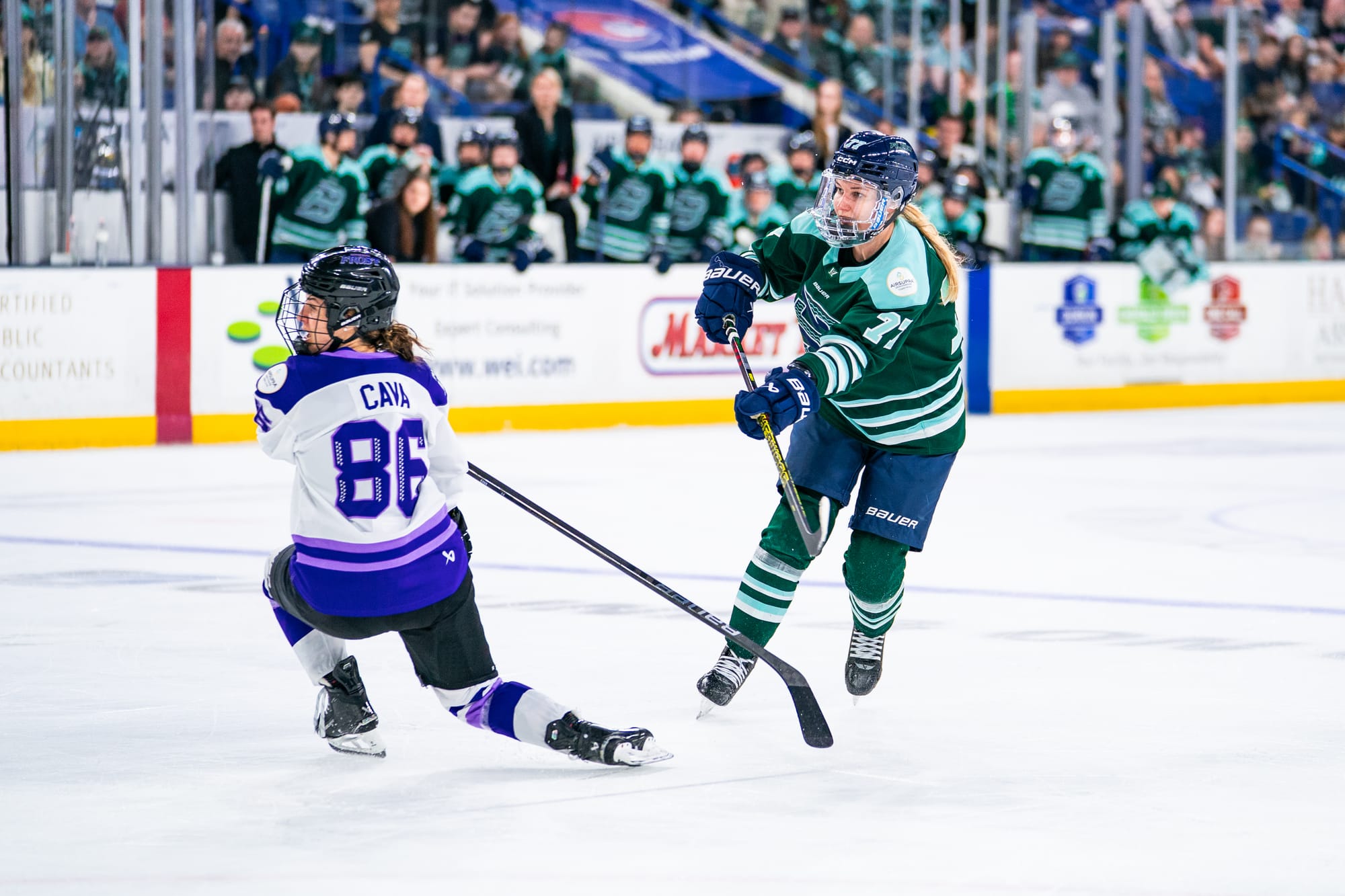 Tapani watches follows through with her stick after playing the puck. Michela Cava is down on one knee in front of her after trying to block the play. Tapani is in a green home uniform, while Cava is in white.