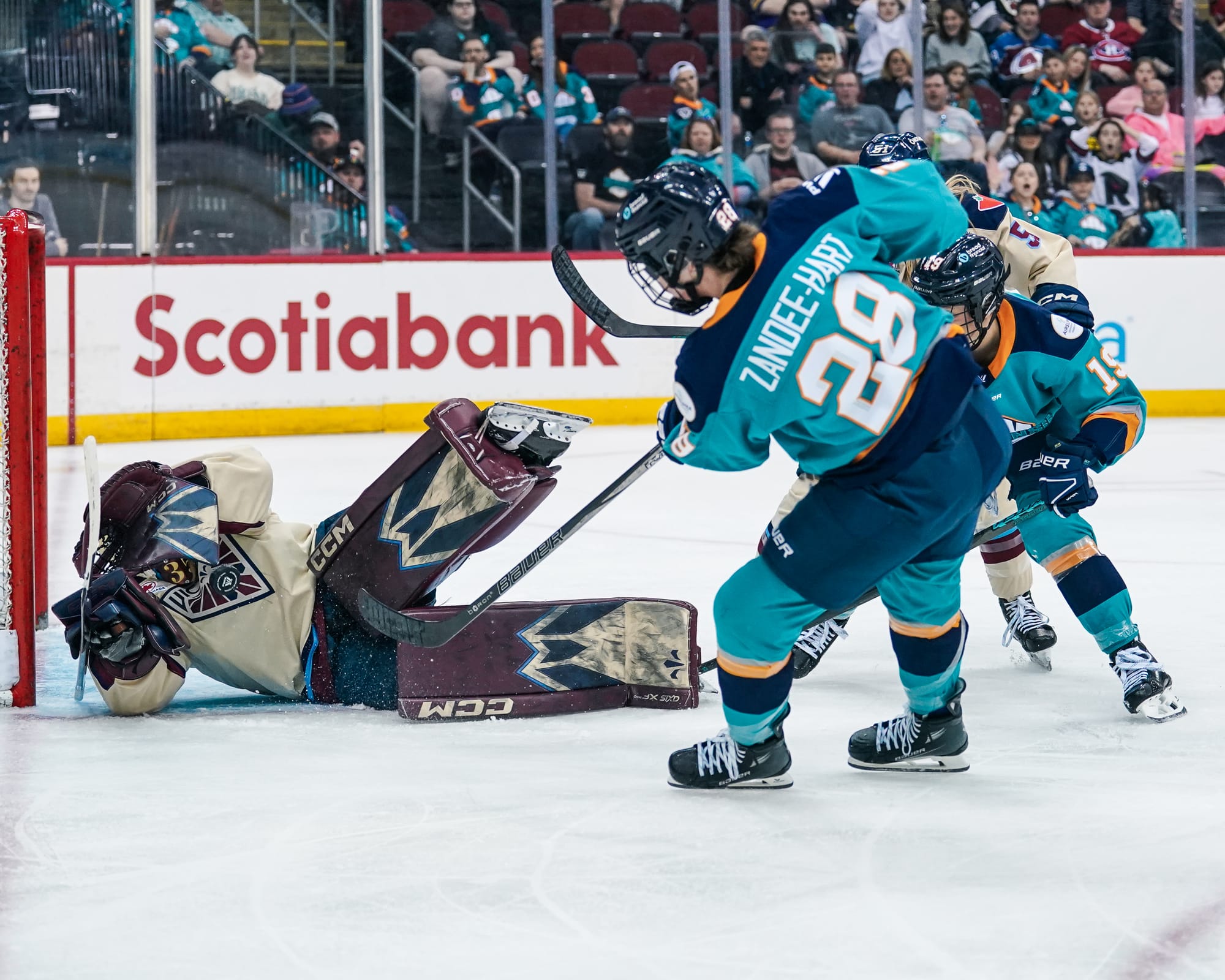 Zandee-Hart leans to her left as she follows through with her stick after shoveling the puck on goal. She is just outside Ann-Renée Desbiens crease, and Desbiens is on the ground after making the save with her hands shielding her mask. Zandee-Hart is in a teal home uniform, while Desbiens is in cream.