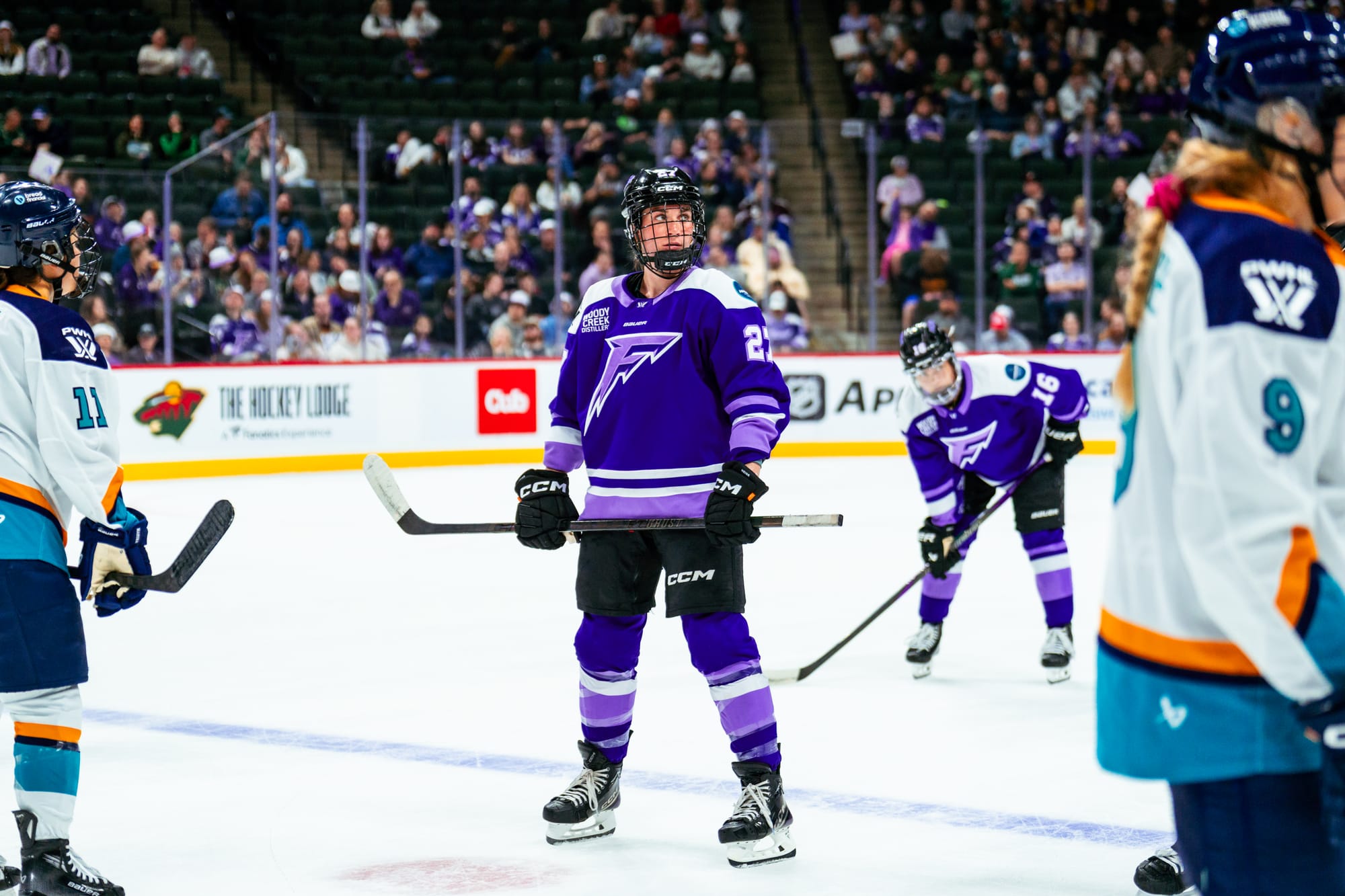 Heise looks to her left as she stands straight up, holding her stick at waist height with two hands. She is wearing a purple home uniform.