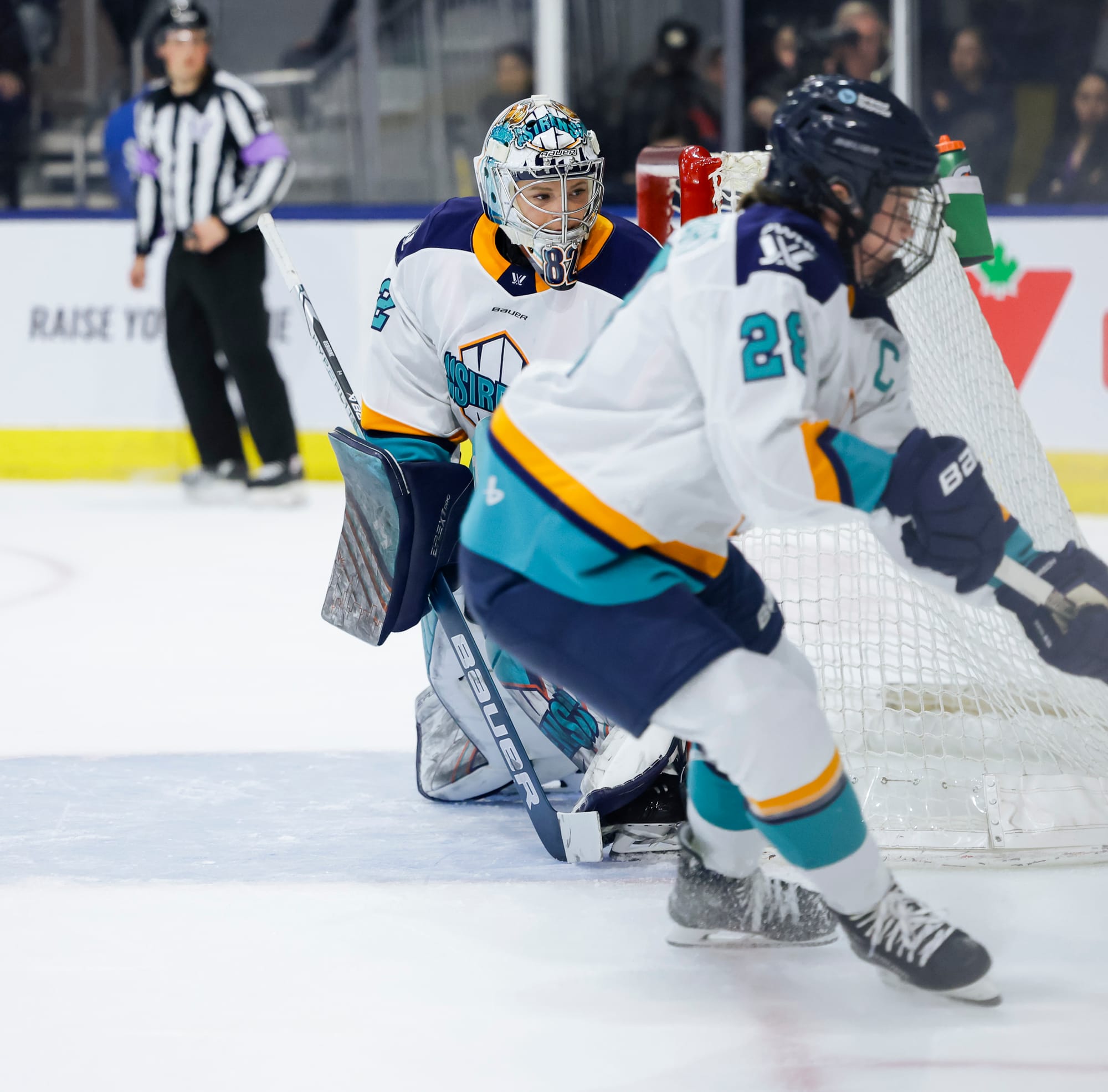 Osborne looks behind the net to her left as Zandee-Hart makes a play on the puck. They are both wearing white away uniforms.