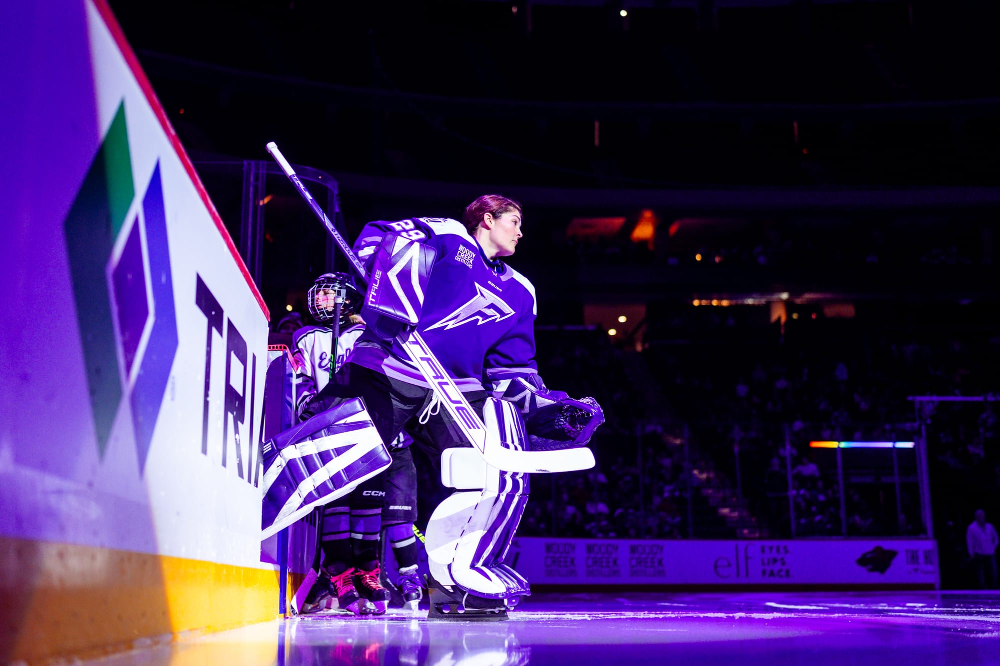 A helmetless Henlsey skates out from the bench onto the ice. She is wearing a purle home uniforms and her purple and white patterned pads.