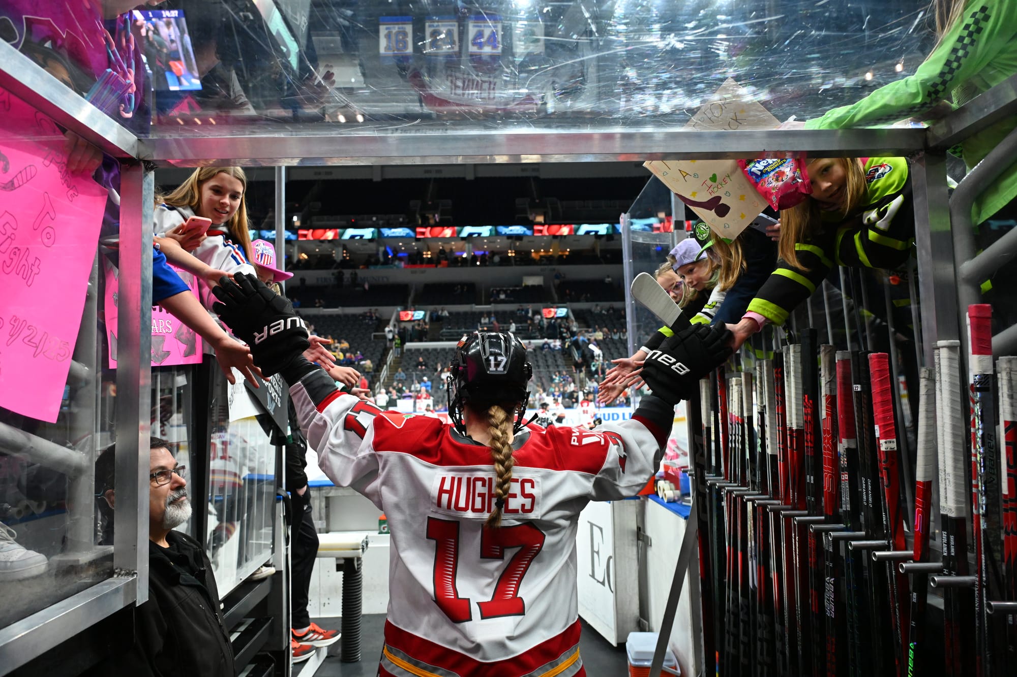 Hughes reaches both arms above her head and out to the sides to fist bump fans as she walks out of the tunnel. The shot is from her waist up and from behind, and she is wearing a white away uniform.