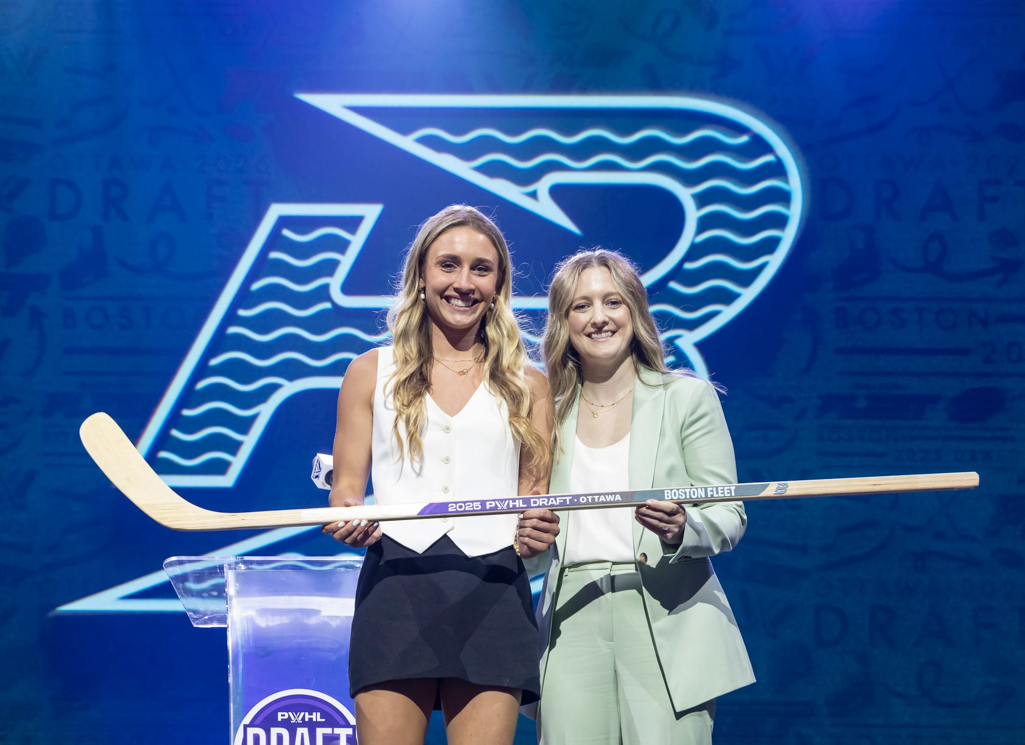 A waist-up shot of Huber and Marmer smiling and holding a wooden stick with "2025 PWHL Draft" and "Boston Fleet" printed on it.