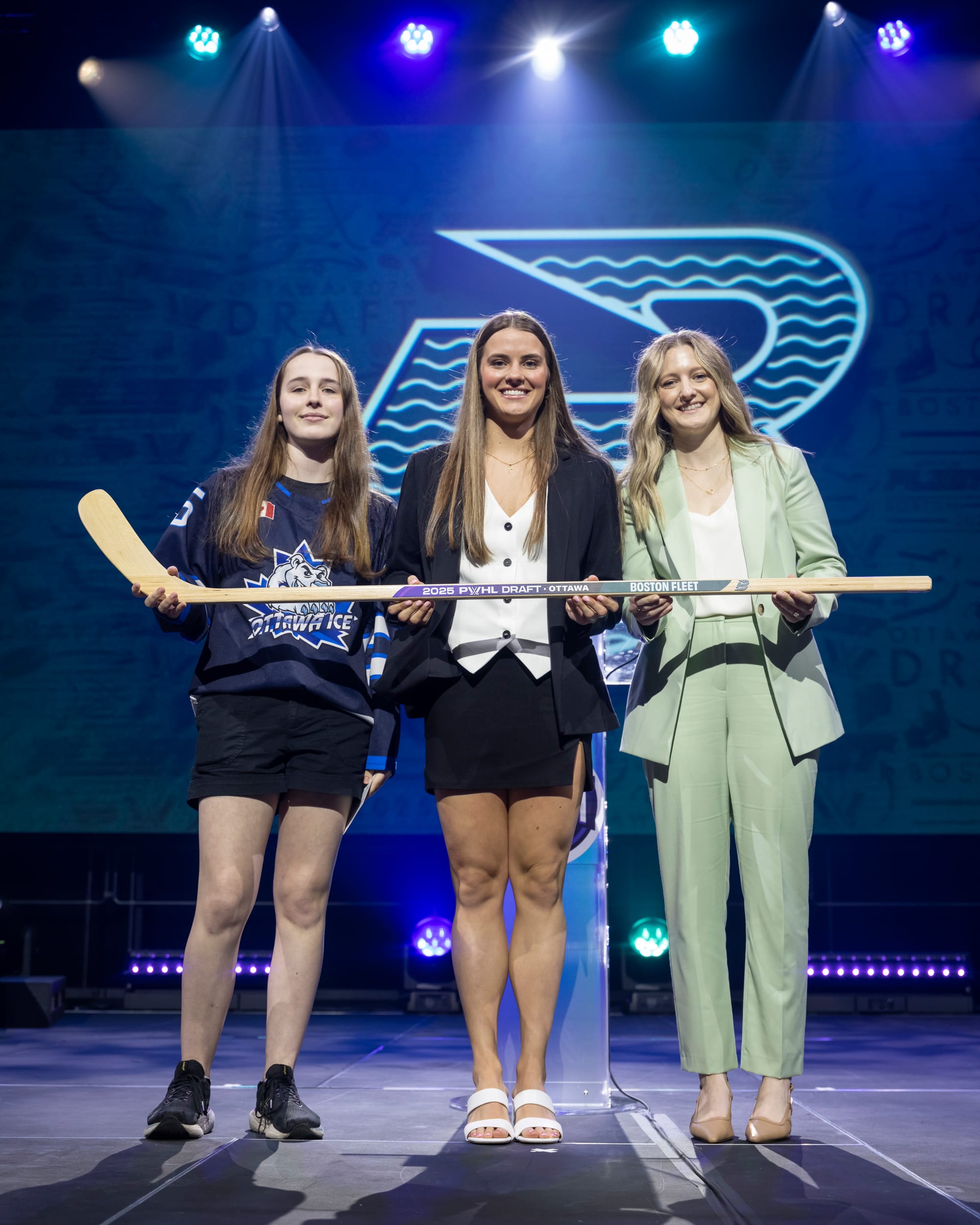 From left to right: the youth player, Brengman, and Marmer smile and hold a wooden stick with "2025 PWHL Draft" and "Boston Fleet" printed on it.
