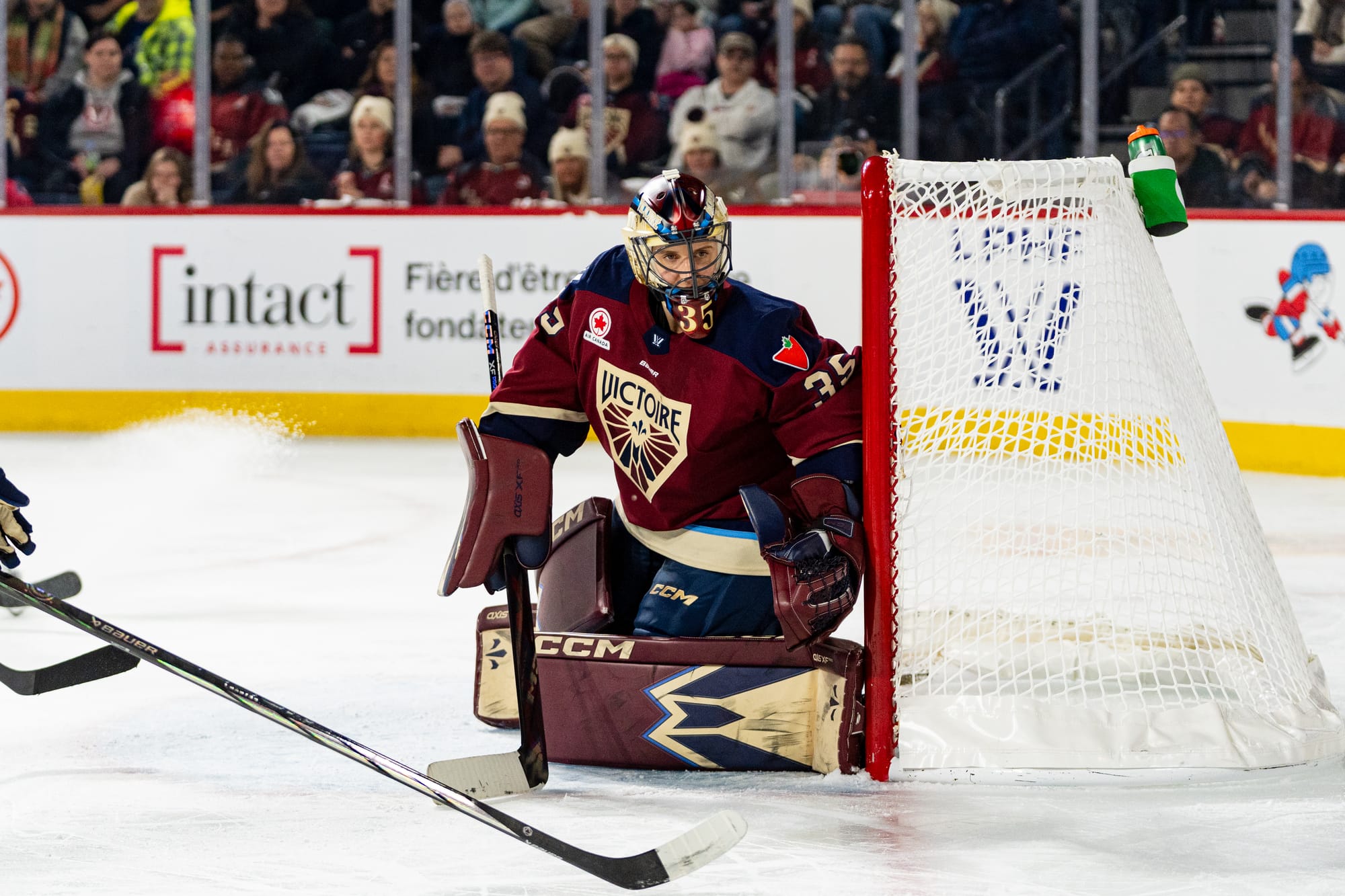 Desbiens looks around her post at the play in the corner, which is out of frame. She is on her knees and wearing a maroon home uniform.