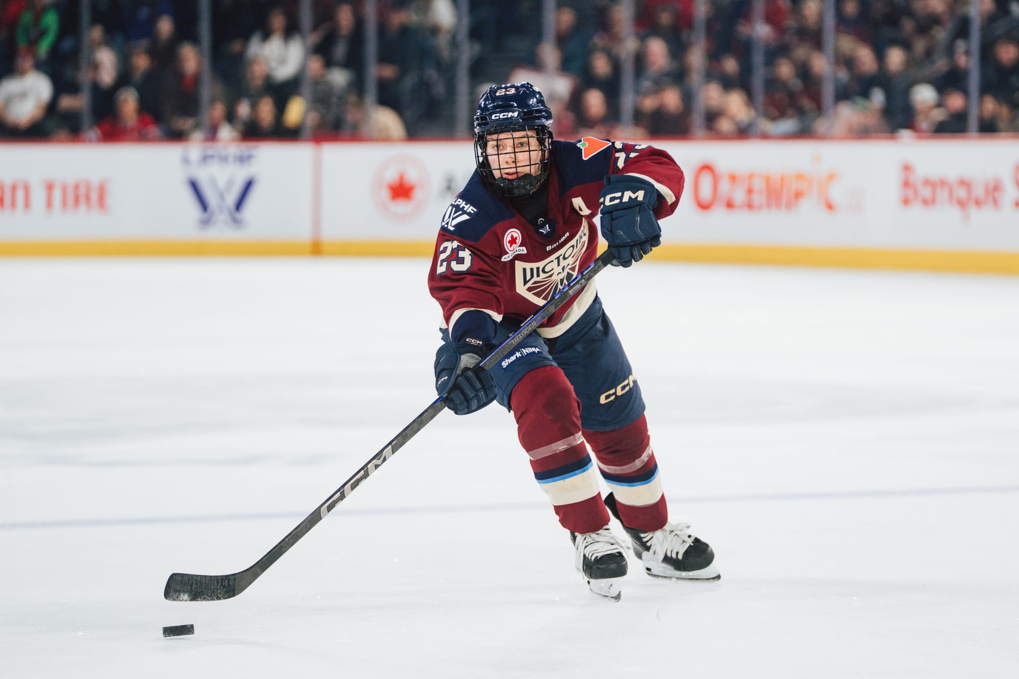 Ambrose skates across a blue line as she passes the puck to an out-of-frame teammate. She is wearing a maroon home uniform.