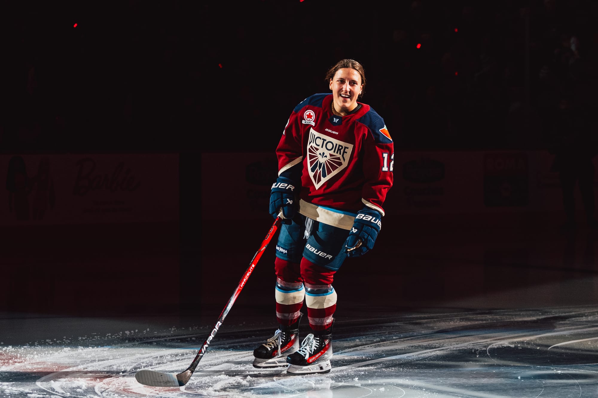 Gardiner skates past center ice under a spotlight, smiling and looking to her left. She is wearing a maroon home uniform. 