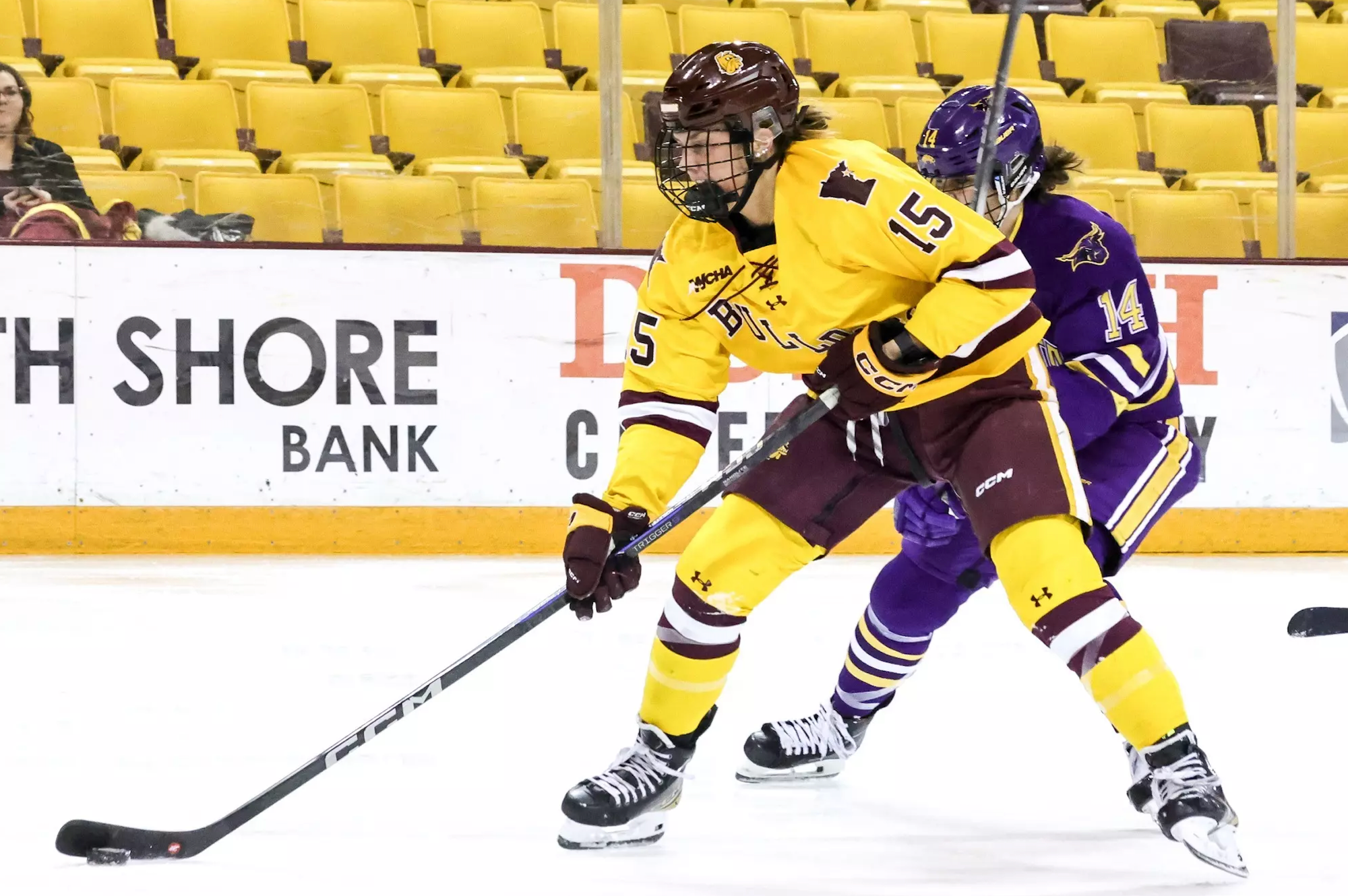 Mobley skates and protects the puck from a defender. She is wearing a gold UMD uniform.