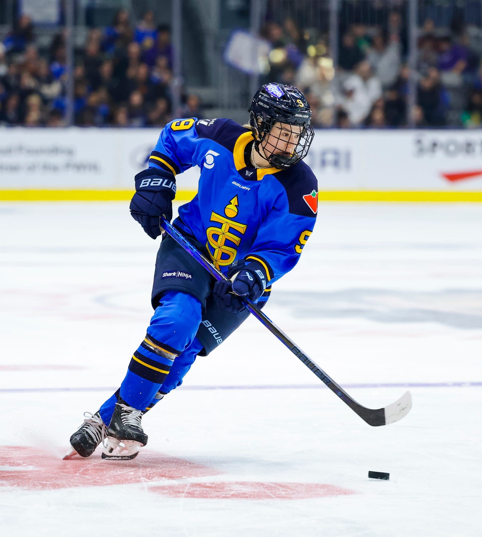 Watts looks down but also ahead of her as she skates with the puck. She is wearing a blue home uniform.