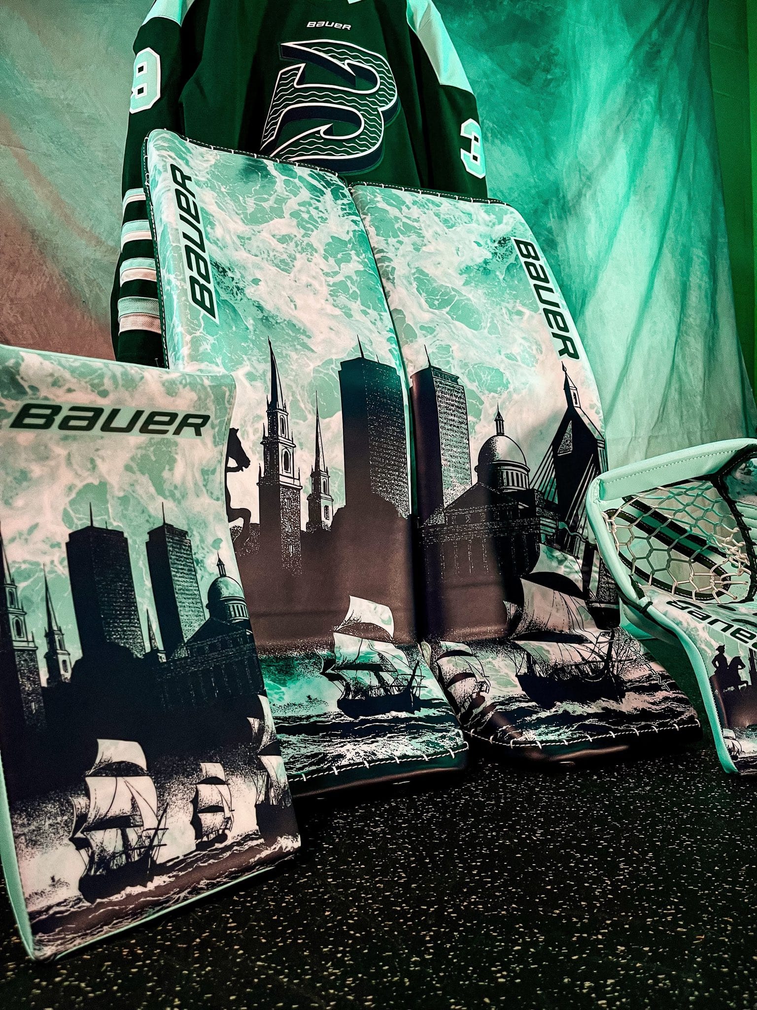 An up-close look of Levy's pads. Several iconic Boston buildings grace the fear, with ships sailing along the bottom. The background is designed to look like water. The drawn designs are black, while the background is the Fleet's teal color.