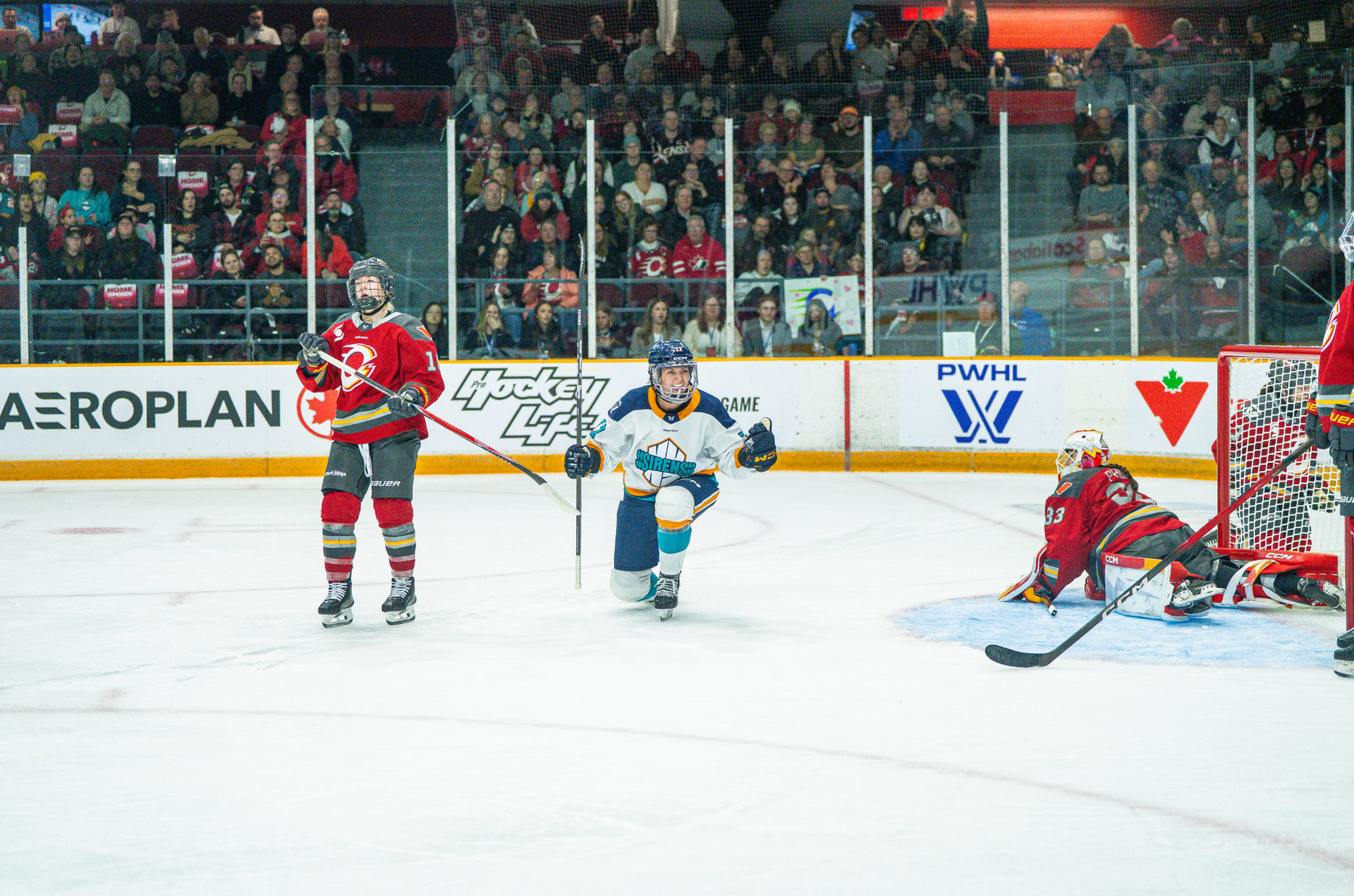 Girard pumps her fists while one knee, smiling as she celebrates one of her three goals. She is wearing a white away uniform.