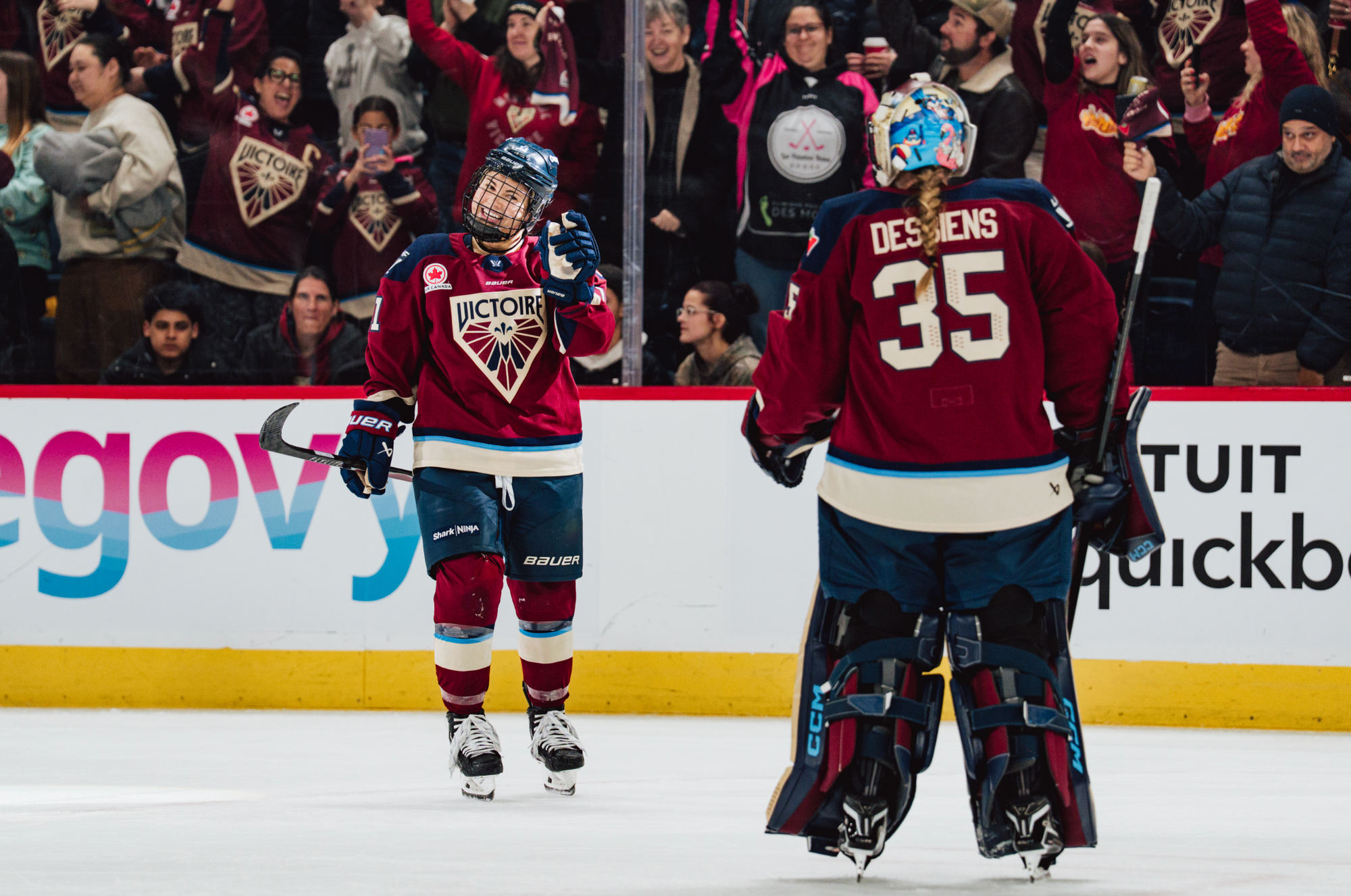 Roque, facing the camera, smiles wide and points at Desbiens, who has her back to the camera. The crowd celebrates alongside them in the background. Both players are wearing maroon home jerseys.