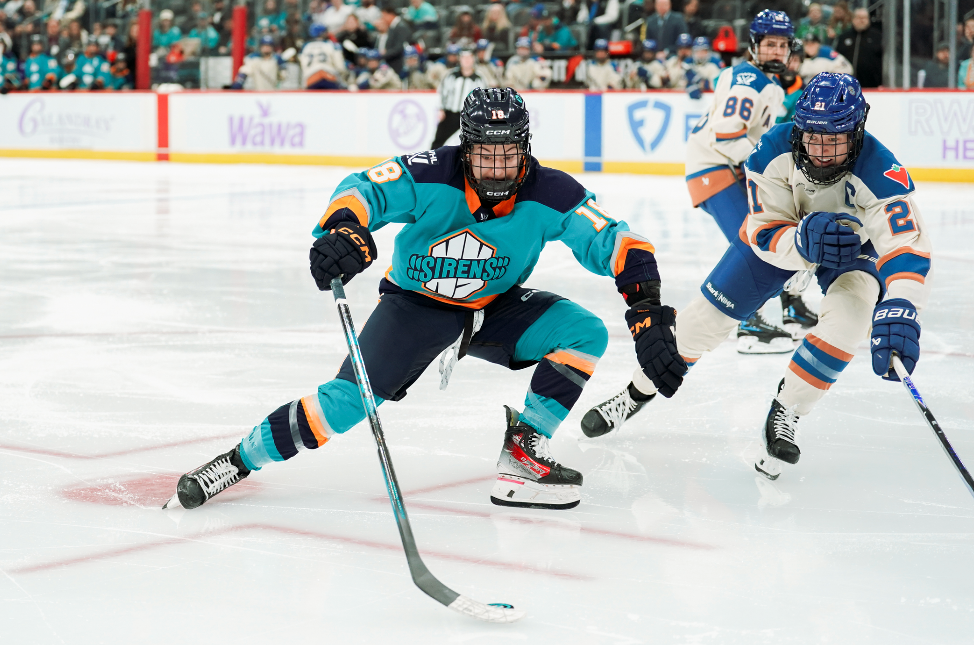 Wheeler uses her stick to keep the puck away from her body to shield it from the oncoming Bell. Wheeler is in a teal home jersey, while Bell is in a cream away one.
