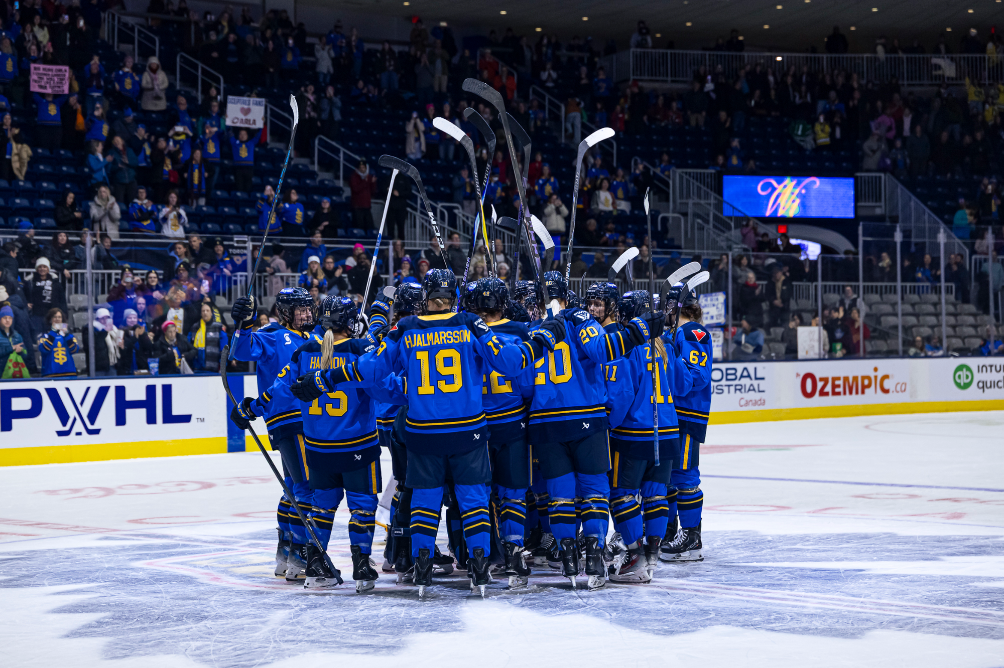 The Sceptres celebrate their first home win of the season with a group hug at center ice. They are all wearing blue home uniforms.