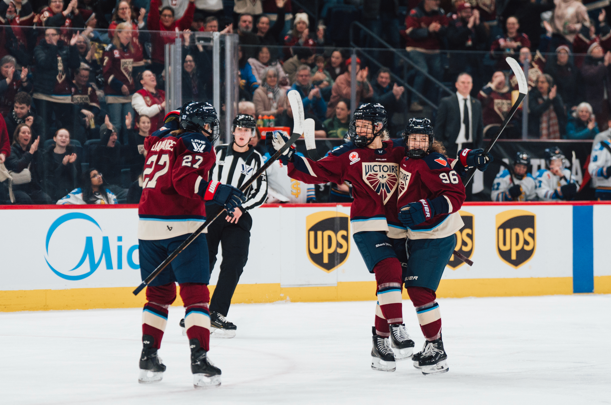 Three Victoire players celebrate a goal with a group hug. They are all wearing maroon home uniforms.